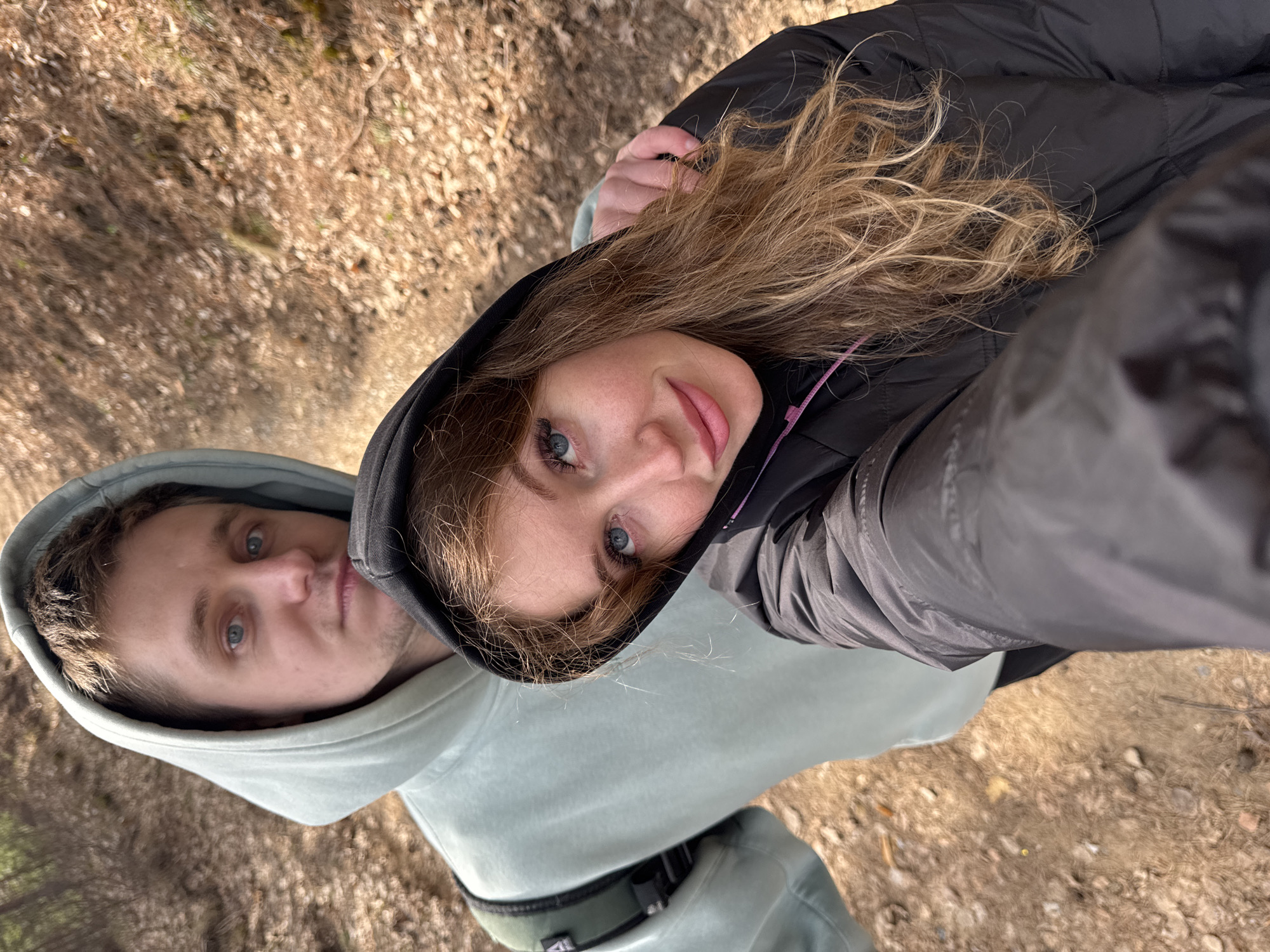A rotated outdoor selfie captures a woman and a man. The woman, in the foreground, has blonde, wavy hair, blue eyes, and a subtle nose piercing, looking towards the camera with a soft smile. She wears a dark hooded jacket. Behind her and slightly to her left is a man with short light brown hair and blue eyes, wearing a light grey-blue hooded sweatshirt, his expression calm.
The setting is an outdoor environment, likely a forest or wooded area, with the ground covered in dry leaves, twigs, and dirt, suggesting autumn or early spring. The background is blurred, showing more of the natural ground texture. The lighting is bright and natural, indicating a sunny day. Their clothing suggests cool but not cold weather. The overall mood is relaxed, implying they are on a walk or hike in Noviy, Russia. No text is visible in the image.
