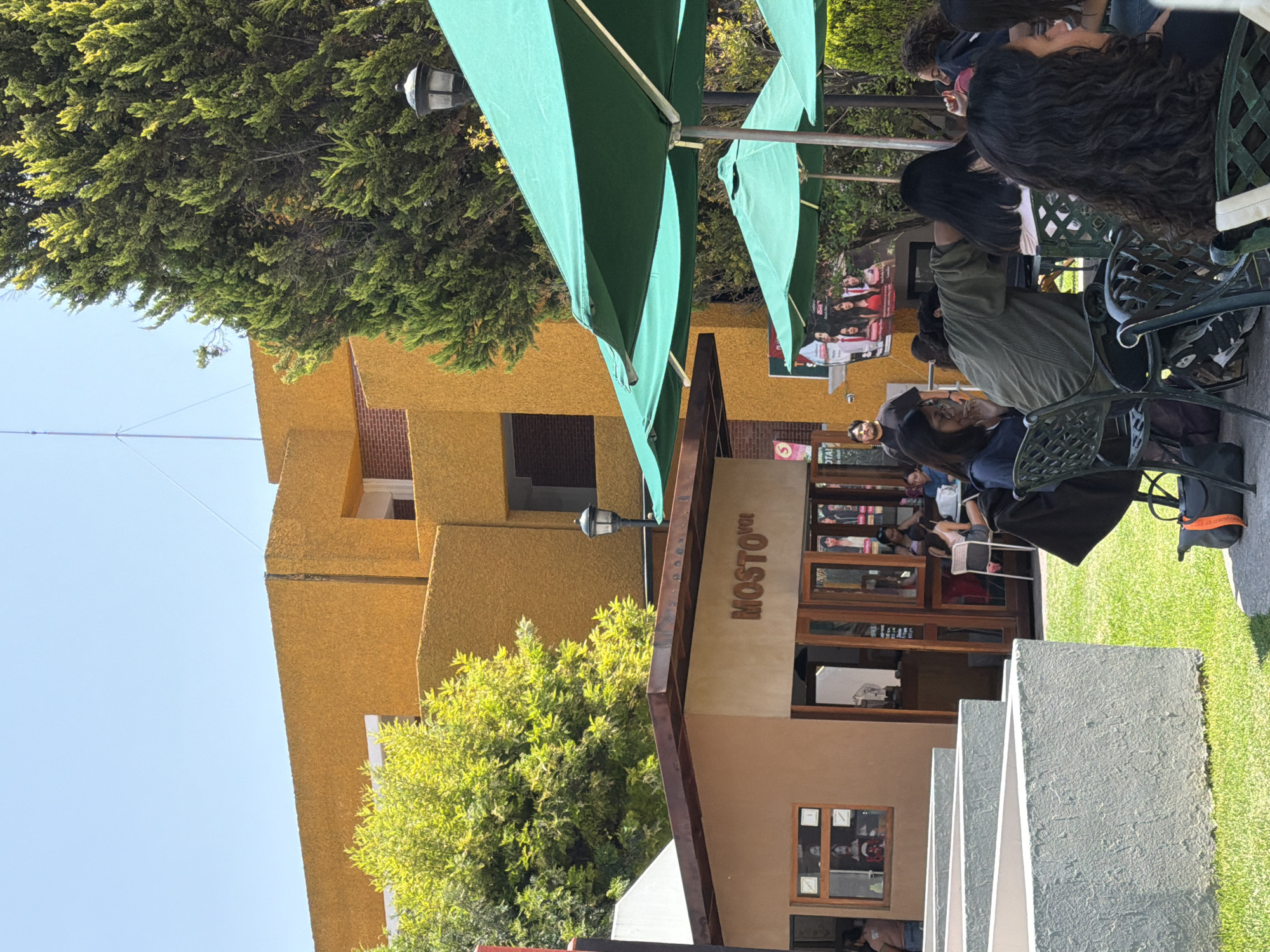 The image captures an outdoor scene with a distinctive yellow building in the background, likely made of concrete. The building's architecture features stepped or tiered sections. In front of the building, there is a cafe or restaurant named "MOSTO". Several people are seated at tables with green umbrellas, suggesting an outdoor seating area. Patrons are engaged in conversations and eating. The scene appears to be in a city with tree coverage. Steps and some greenery are visible in the foreground, indicating an outdoor setting.