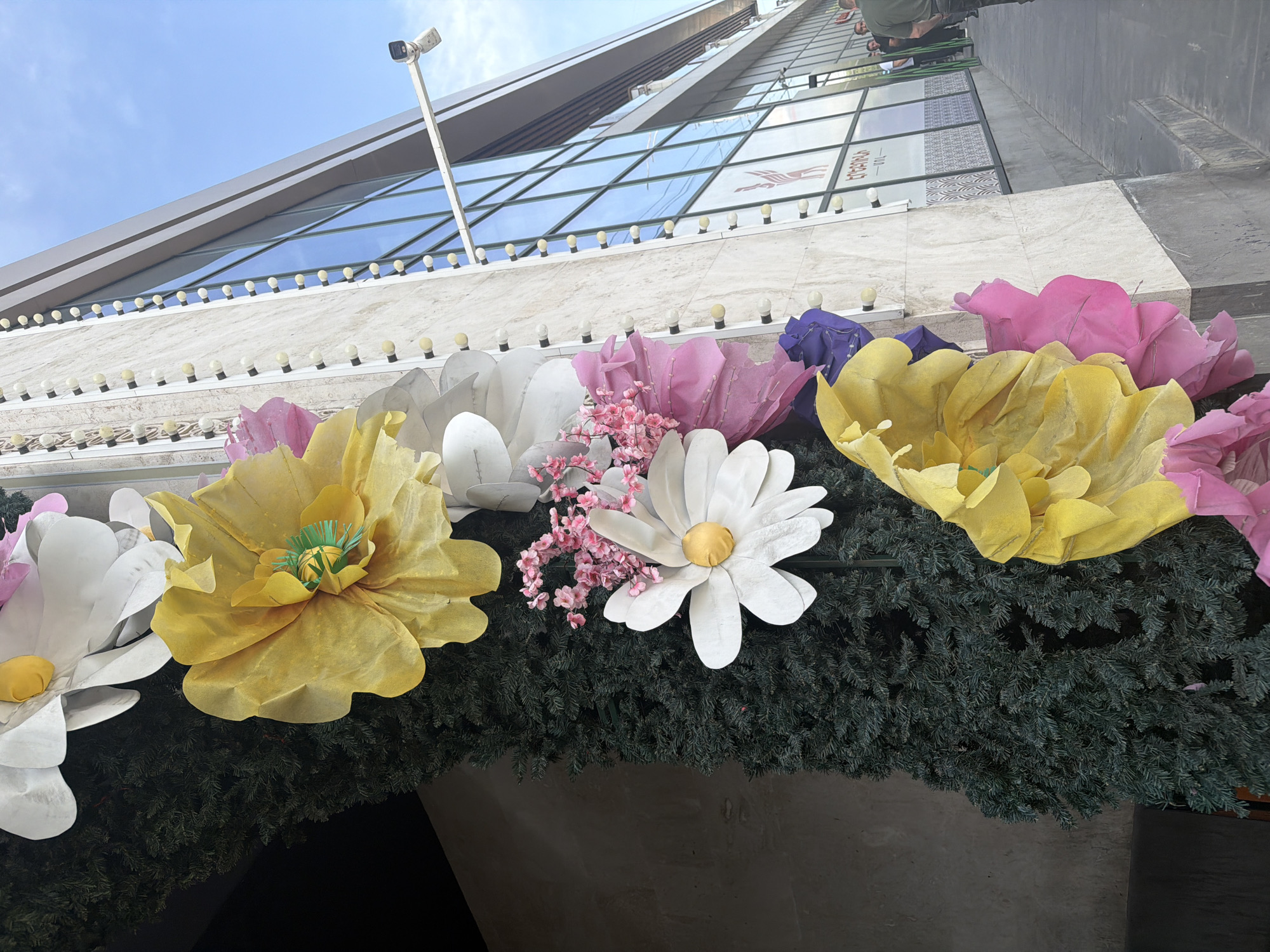 The image captures a low-angle view of decorative floral arrangements juxtaposed against a modern architectural facade, likely in an urban setting. Large, stylized paper flowers in shades of white, yellow, and pink are prominently displayed, with a smaller cluster of pink blossoms interspersed among them. These decorations sit atop a dense, evergreen garland.
In the background, a multi-story building with a reflective glass exterior rises into the sky. A surveillance camera is affixed to a pole that is also adjacent to the building. A row of small, globe-shaped lights runs along the edge of the building's structure.
At the very top of the frame, a small portion of a person or people is visible.
Although Yerevan, Armenia is requested, this information cannot be verified based on the visual data provided in the image.