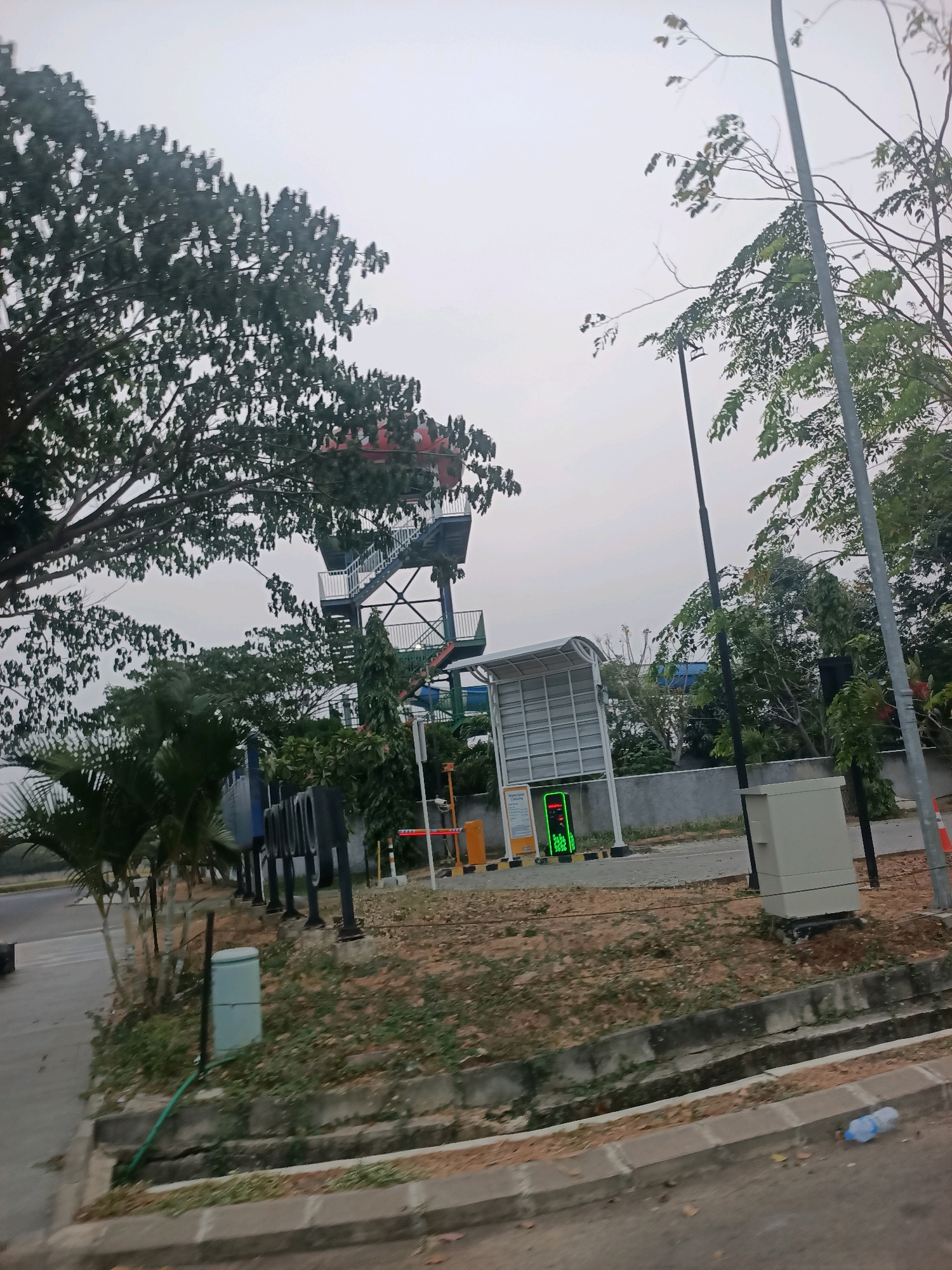 This is a medium-distance, eye-level shot of a water park entrance area on an overcast day. In the background, a tall, multi-story water slide structure is visible, with a red top section and metal stairs. To the right of the slide, a white, modern-looking structure with a curved roof covers a ticketing or information booth. A bright green digital display is mounted on a yellow barrier gate, likely indicating the operational status of the park or a charging station. Several large trees with lush green foliage are prominent in the mid-ground and left side of the image, with palm trees and other smaller vegetation in the foreground. A concrete sidewalk and drainage ditch run along the bottom of the image, with a discarded plastic water bottle on the sidewalk. Several dark, upright poles with curved tops are lined up along the left side of the scene, possibly part of a parking meter system or decorative bollards. A light-colored utility box is situated on the right side of the image. The overall impression is of a recreational facility entrance in an urban or suburban environment. The image appears to have been taken from a moving vehicle, given the slight blur and angle.