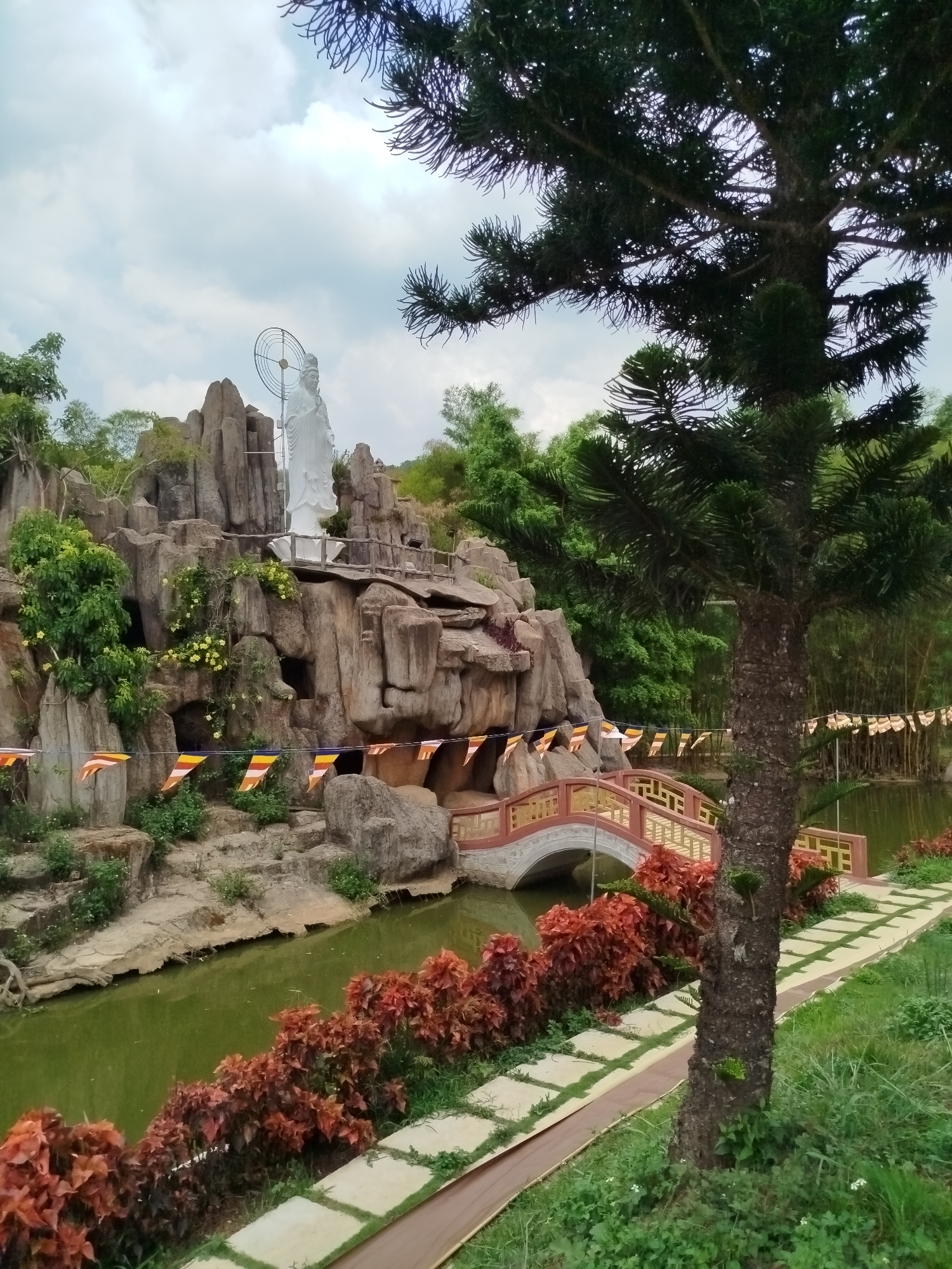 The image captures a tranquil scene in Da Lat, Vietnam, featuring a Buddhist temple complex. A white statue of a deity stands prominently atop a rocky outcrop, overlooking a small pond. The landscape includes lush greenery, including a variety of trees and shrubs. A decorative bridge with red and gold accents spans the water. A paved pathway runs along the edge of the pond, bordered by red foliage. Buddhist prayer flags are visible, strung along the rocks and the bridge. The overall atmosphere suggests a peaceful, contemplative environment.