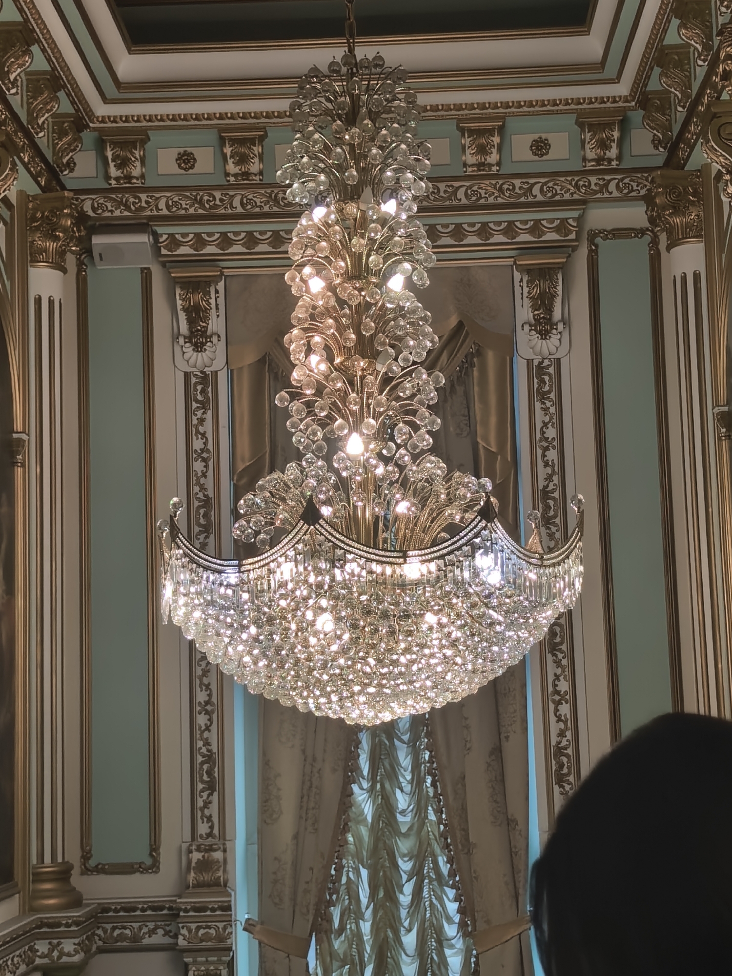 An ornate, multi-tiered crystal chandelier hangs from a decorated ceiling in a dimly lit room with ornate gold trim and pale green walls. The chandelier is the central focus, with numerous clear crystals reflecting light from within, creating a dazzling effect. The ceiling features recessed panels and gold molding. On either side of the chandelier, gold-detailed columns and decorative wall panels are visible. Heavy, patterned curtains are drawn behind the chandelier, suggesting a window or alcove. The bottom right corner of the image is obscured by the dark silhouette of a person's head and shoulders.