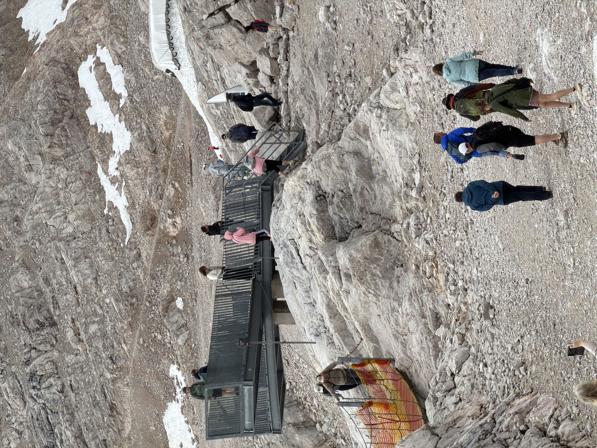 The image depicts a mountainous, rocky terrain near Garmisch-Partenkirchen, Germany, likely at the Zugspitze summit or a similar high-altitude attraction. A group of people are engaged in various activities, including walking on pathways and stairs, standing on elevated platforms, and taking photographs.
In the foreground, several individuals are walking away from the viewer on a gravel path. They are dressed in casual outdoor clothing, with backpacks visible on some. To the left, a person is ascending a set of stairs leading to an elevated metal walkway. This walkway, with railings, is occupied by several other individuals observing the surroundings. One person in a pink hooded jacket is descending a set of stairs from this walkway. Further in the background, more people are visible on the rocky slopes and near a white tent-like structure. The environment is characterized by barren rock, patches of snow, and a rugged, uneven landscape. A woman in the lower right corner is holding up a mobile phone, appearing to take a photo. The overall atmosphere suggests a tourist destination where visitors are exploring the natural scenery.