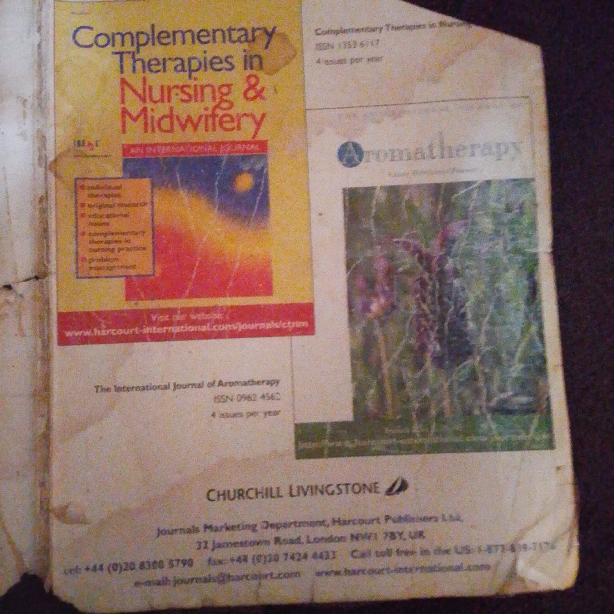 The image displays the front cover of a journal titled "Complementary Therapies in Nursing & Midwifery" alongside a section promoting "Aromatherapy". The journal cover features the title in large purple and red lettering against a yellow background, with smaller text providing publication details and subscription information. It also includes bullet points listing the types of content found in the journal. Below this section, a website address is visible.
To the right, the "Aromatherapy" section shows a logo and text, with a photographic background of what appears to be lavender or a similar plant.
At the bottom of the image, there is a publisher's imprint for "CHURCHILL LIVINGSTONE", followed by contact details for "Journals Marketing Department, Harcourt Publishers Ltd." This includes an address in London, UK, a phone number, and a website.
The image is a photograph of the journal, likely taken indoors under artificial lighting, as suggested by the shadows and the overall color balance. The quality of the image is somewhat degraded, with visible graininess and discoloration, particularly around the edges, indicating the age or condition of the physical item being photographed. There are no visible signs or indications related to the city of Jalingo, Nigeria.