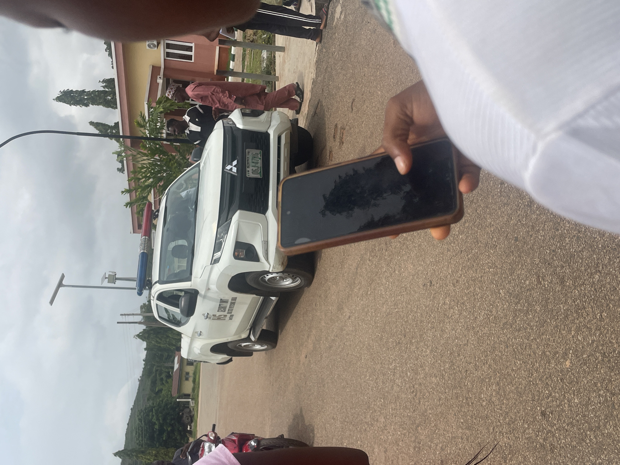 A white Mitsubishi L200 pickup truck with flashing blue and red lights is parked on an asphalt road in Lokoja, Nigeria. The truck has "SECURITY UNIT" written on its side. Two men are standing near the truck, and another man is walking away from it. In the foreground, a person's hand is holding a smartphone, partially obscuring their face. In the background, there is a building with a peach-colored facade, trees, and a cloudy sky. A red motorcycle is parked to the left of the truck. The environment appears to be an outdoor setting within a built-up area.