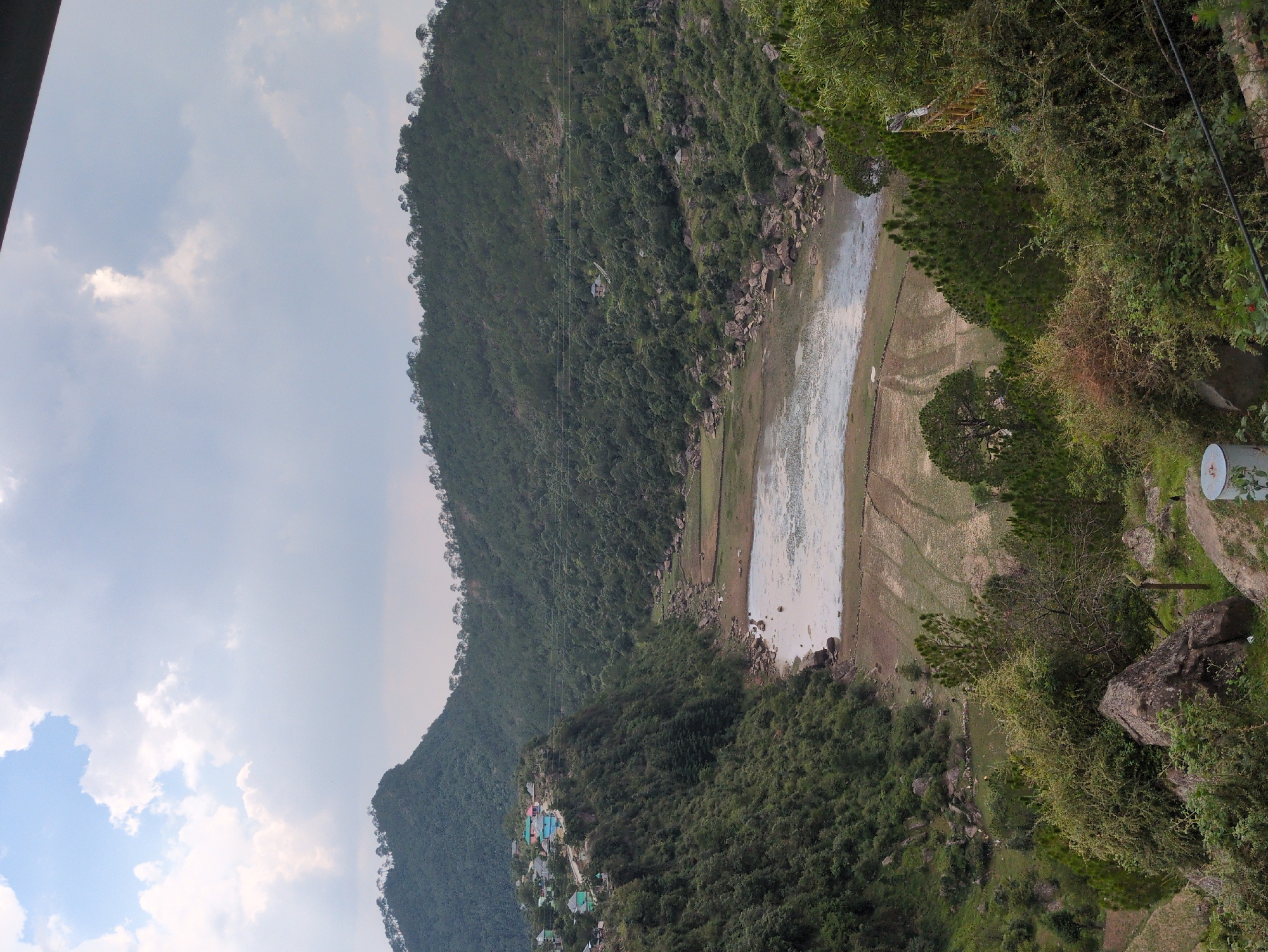 The image shows a panoramic view of a valley with a lake in the center, surrounded by lush green hills. The hills are densely covered with trees and vegetation. On the left side of the valley, a small village with colorful houses is perched on a hillside. In the foreground, there are rocky outcrops and dense foliage, including pine trees. The sky is partly cloudy. The scene depicts a rural landscape with natural beauty. The location is the city of Doh, India.