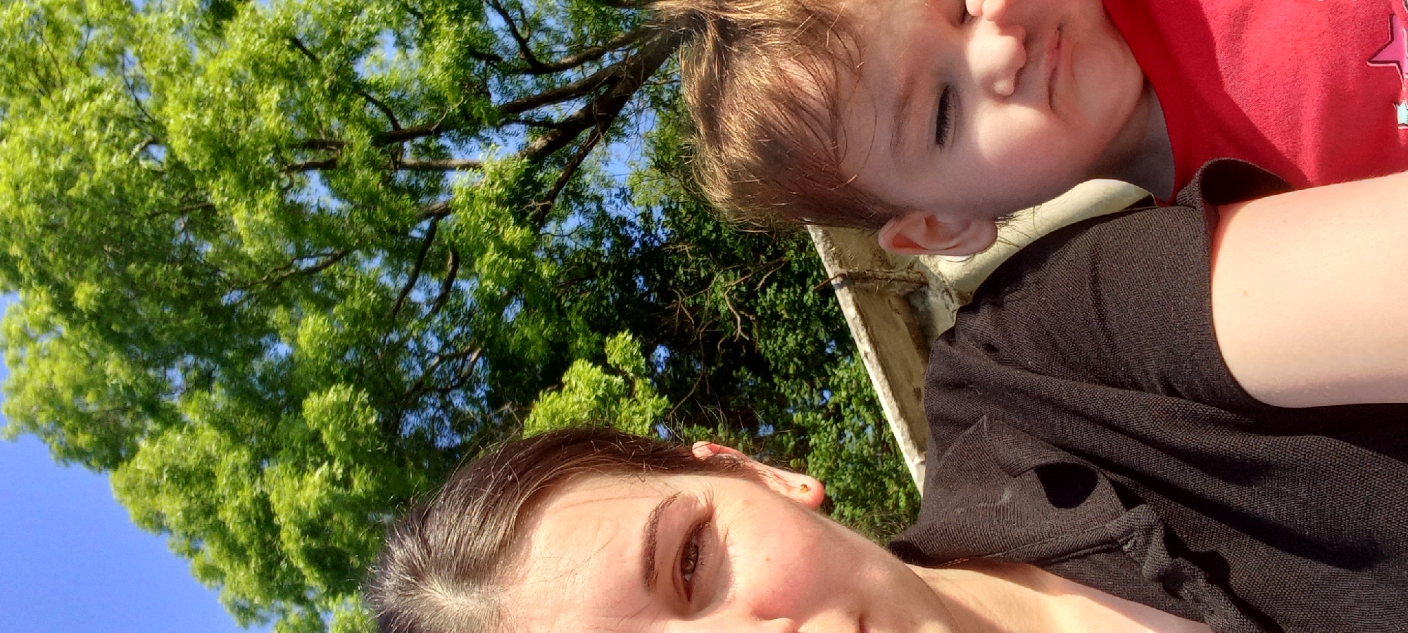 The media file is a low-angle, close-up photograph featuring two individuals outdoors, likely a mother and her child. The dominant elements are the lush green foliage of a tree overhead and the bright blue sky. In the foreground, a person's face is visible, with dark hair and brown eyes looking towards the camera. To the right, partially obscuring the background, is another person, a child, with light brown hair and closed eyes. The child is wearing a red shirt with a star graphic. Both individuals appear to be lying down, with the camera angled upwards from a low vantage point. The overall lighting suggests a sunny day, possibly in the late afternoon or early morning due to the warm tones. The setting appears to be a natural outdoor environment, with the abundance of green leaves indicating a warm season. The relaxed posture of the subjects and the casual nature of the shot suggest a moment of leisure and connection. There is no visible text in the image.