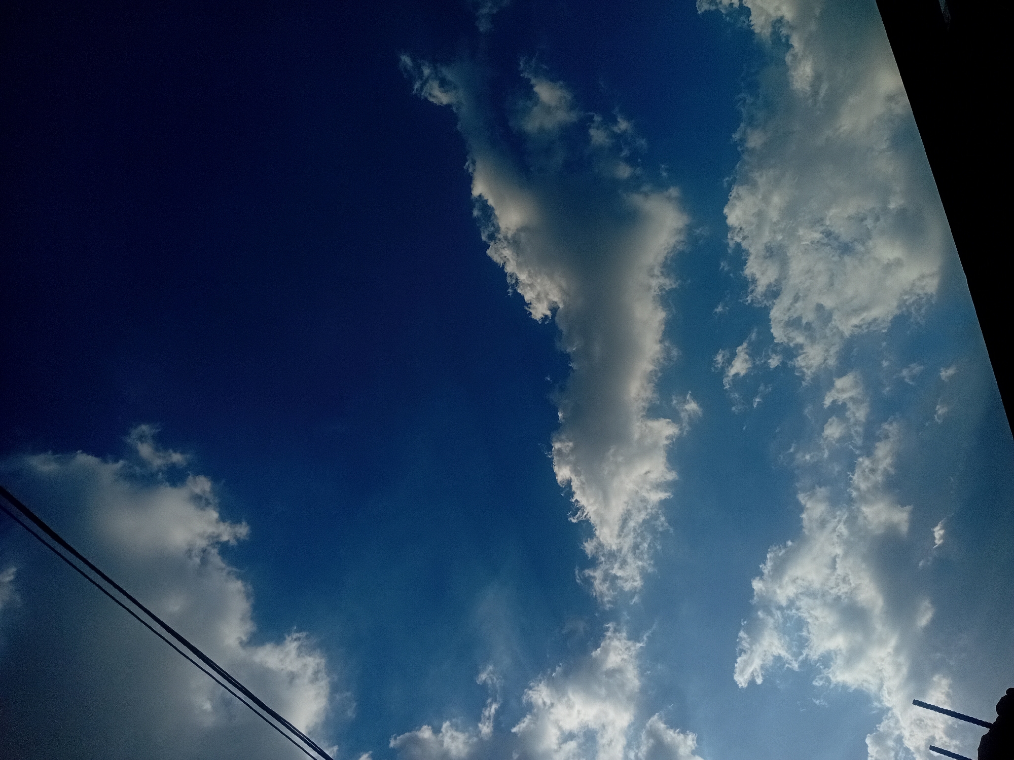 The image displays a clear blue sky interspersed with various cloud formations, captured during daylight hours in Lokoja, Nigeria. The sky exhibits a gradient of blue, transitioning from a darker, deeper shade in the upper left quadrant to a lighter, brighter blue towards the lower and right portions of the frame. White, fluffy clouds, brightly illuminated by sunlight, are scattered across the sky, contrasting with larger, greyish clouds that appear more shadowed and dense.
In the lower-left corner, two dark, parallel lines, likely overhead power lines or utility cables, cut across the scene, silhouetted against the sky and clouds. On the right side, dark, indistinct shapes intrude from the edges, possibly parts of a building, pole, or foliage, creating a partial silhouette against the bright backdrop. No people or specific events are depicted, and there is no visible text. The scene is a static depiction of the sky and atmospheric conditions, characteristic of a partly cloudy day.