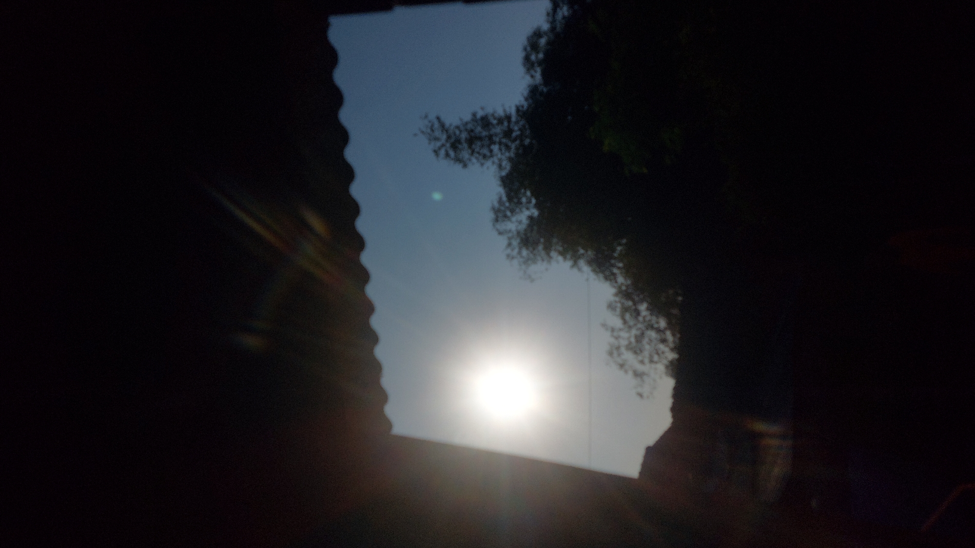 The image captures a view of the sky with the sun visible on the left, casting a bright glow. Silhouetted against the sky are trees and a building. A corrugated roofline is visible at the top of the frame. The scene appears to be outdoors, possibly in a residential or rural area. No specific landmarks indicating the city of Jatirogo, Indonesia, are discernible in the image.