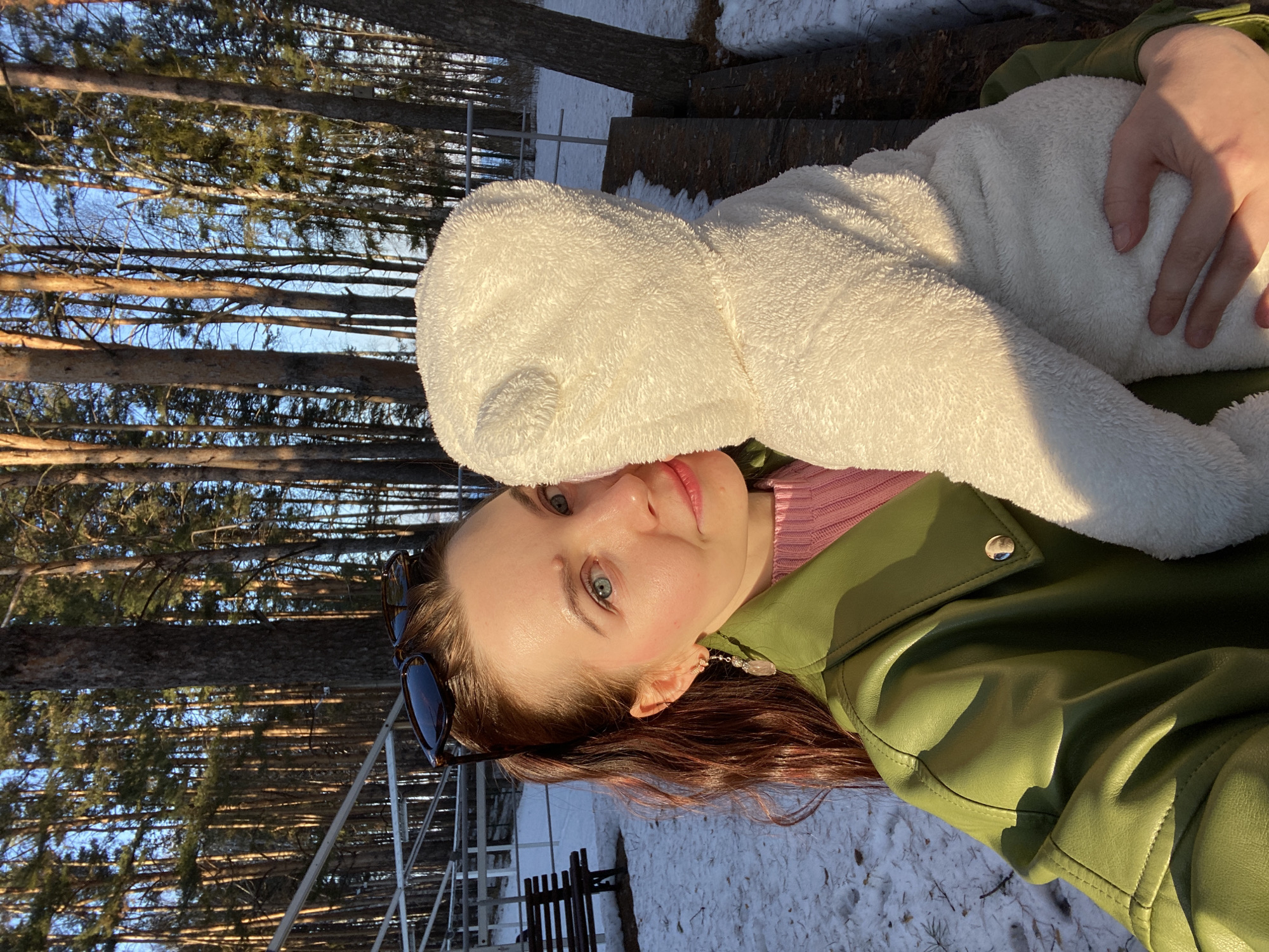 A woman is pictured outdoors, likely in a wooded area in the city of Chaikovsky, Russia, evidenced by the landscape with tall pine trees and snow patches on the ground. She wears a green jacket over a pink top and has a white hooded garment pulled over her head. Sunglasses are visible perched in her hair. She is smiling at the camera. A wooden fence and metal railing are visible in the background. The image is taken in daylight with a clear blue sky visible through the trees.