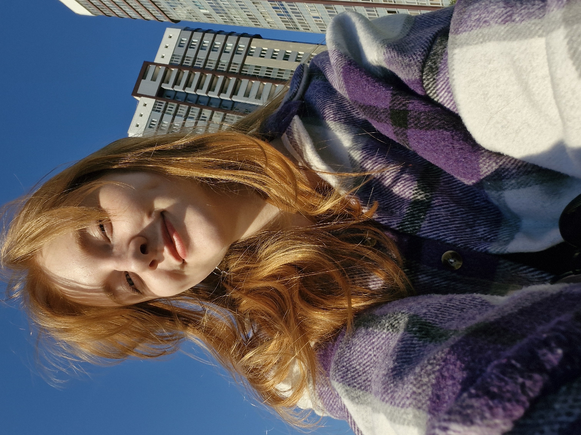 A low-angle close-up shot depicts a fair-skinned woman with long, wavy auburn hair. She is wearing a plaid shirt with shades of white, purple, and black, and it appears to be buttoned. The sky is a clear, bright blue and a tall building with a multi-story, modern design and numerous windows is visible in the background. Based on the architectural style and location, the building appears to be located in Yekaterinburg, Russia.