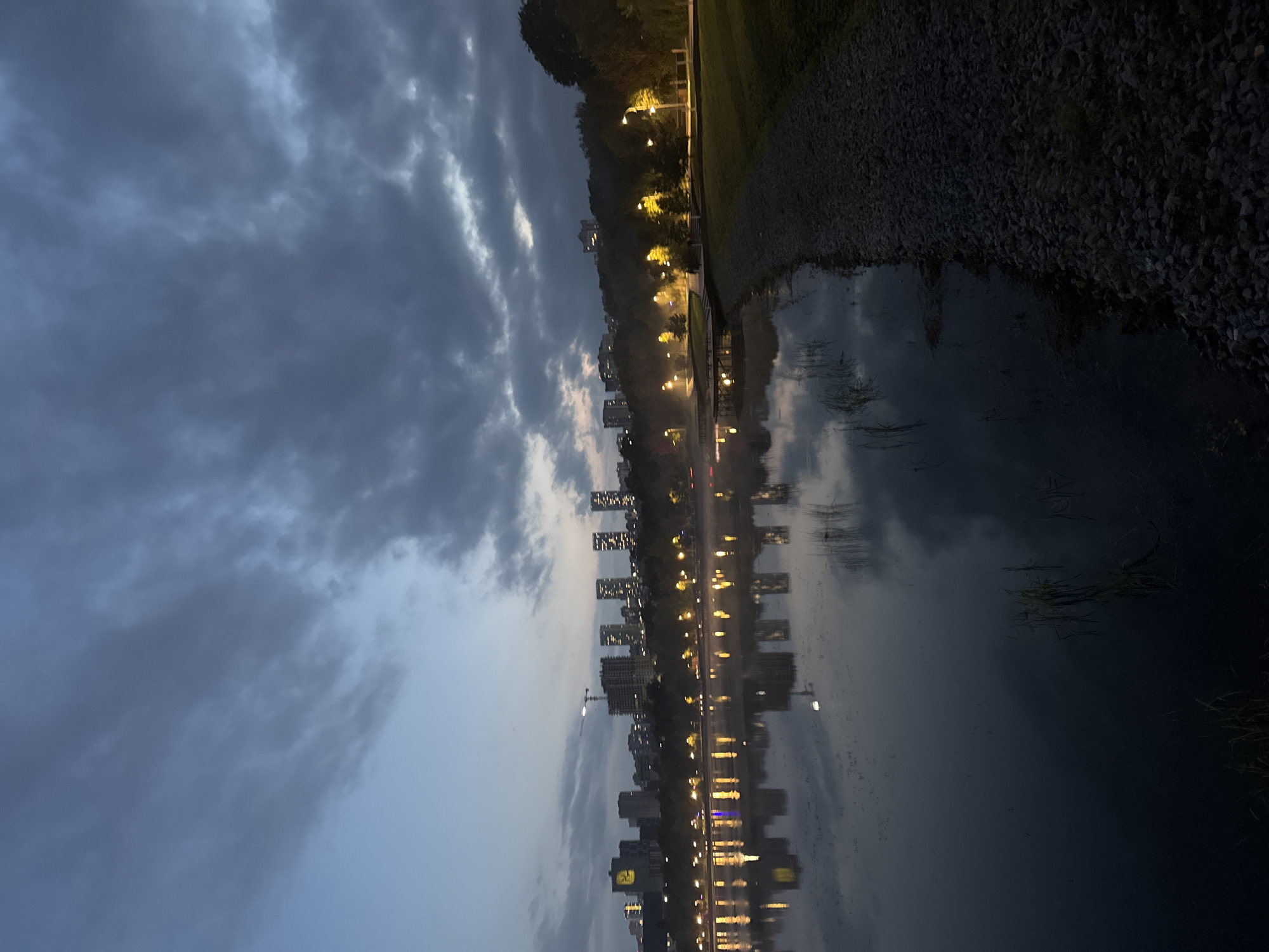 The image depicts a tranquil scene at dusk by a lake, likely in Ufa, Russia, given the presence of apartment buildings and a crane suggestive of ongoing construction. The sky is overcast with dark, dramatic clouds. The water's surface is calm, reflecting the city lights that line the opposite shore and the silhouettes of several tall buildings. A pathway made of gravel runs along the right side of the foreground, bordered by grass and trees. The overall atmosphere is serene, with a touch of urban energy from the distant city lights. No people are visible in the frame.