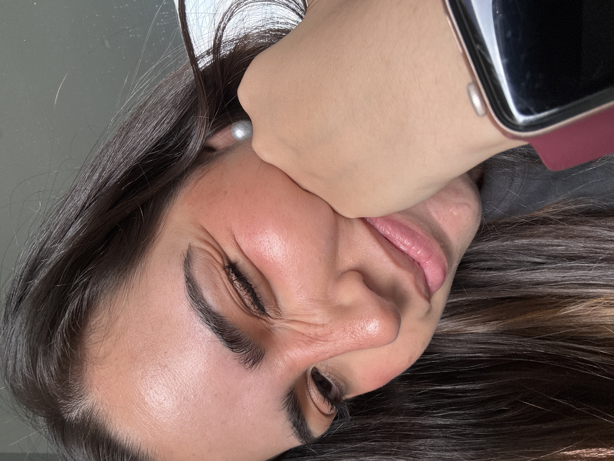 A close-up, rotated portrait photograph depicts a woman with her eyes closed, grimacing as a fist presses against her cheek. She has long, dark hair with highlights and is wearing a pearl earring. A smartwatch with a dark screen and a rose-colored band is visible on the wrist of the hand applying pressure. The background appears to be a neutral gray tone. There is no information available in the image to place it specifically in San Bernardino Tlaxcalancingo, Mexico.