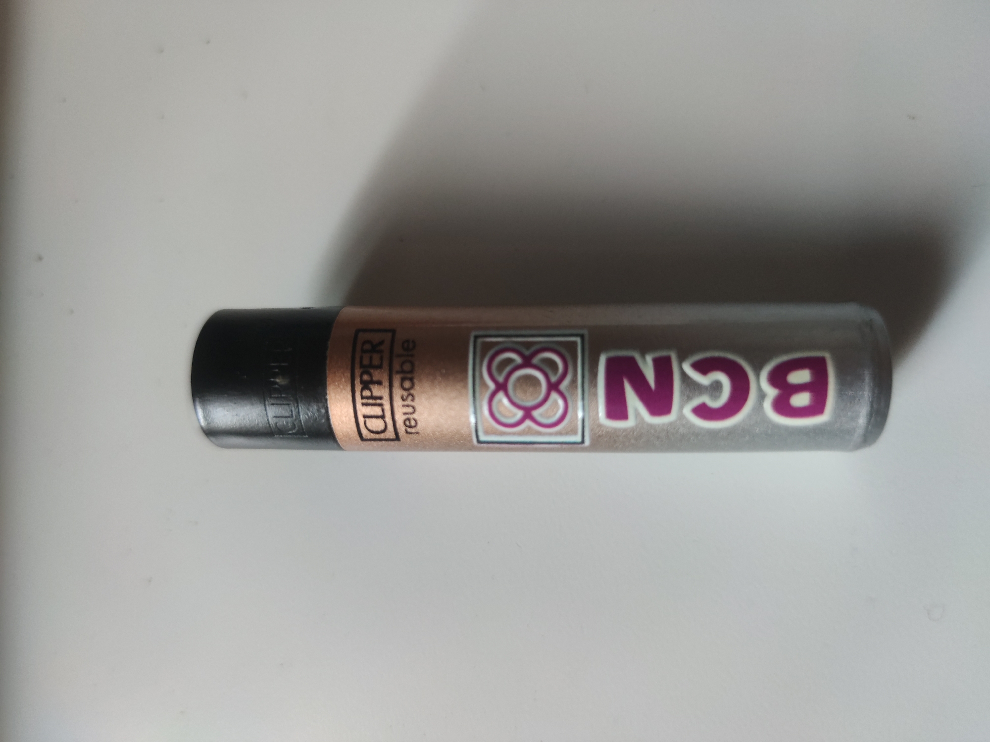 A close-up, top-down shot of a Clipper brand lighter lying horizontally on a white surface. The lighter is cylindrical with a black top. The body is predominantly copper in color, with text that reads "CLIPPER reusable" vertically printed. Adjacent to the text is a square design, featuring an eight-petal flower-like symbol. To the right of the symbol, the letters "BCN" are displayed in a large, bold font. The letters are a metallic silver color outlined in purple. BCN is the abbreviation of the city of Barcelona in Catalonia, Spain.