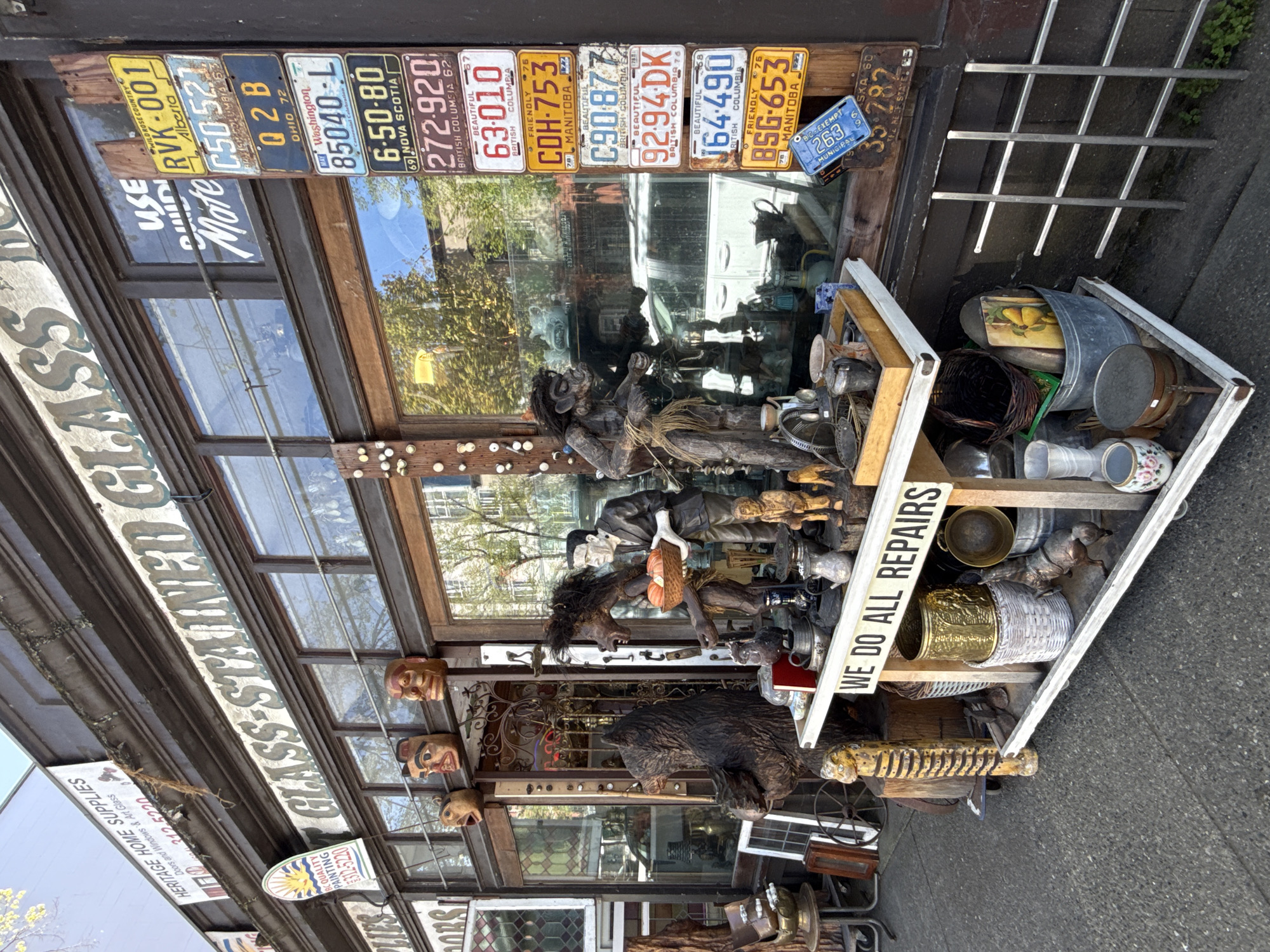 The image shows the facade of a shop in Vancouver, Canada. The shop's name appears to be "Heritage Home Supplies," specializing in doors, windows, and art glass. Above the storefront windows, a collection of license plates from various provinces and states is displayed. The shop also offers glass and stained glass repairs, as indicated by a sign. The shop window displays a variety of items, including wooden carvings of figures and a sign that reads "WE DO ALL REPAIRS." There are also various vintage items such as buckets, baskets, and metal containers displayed outside the shop.