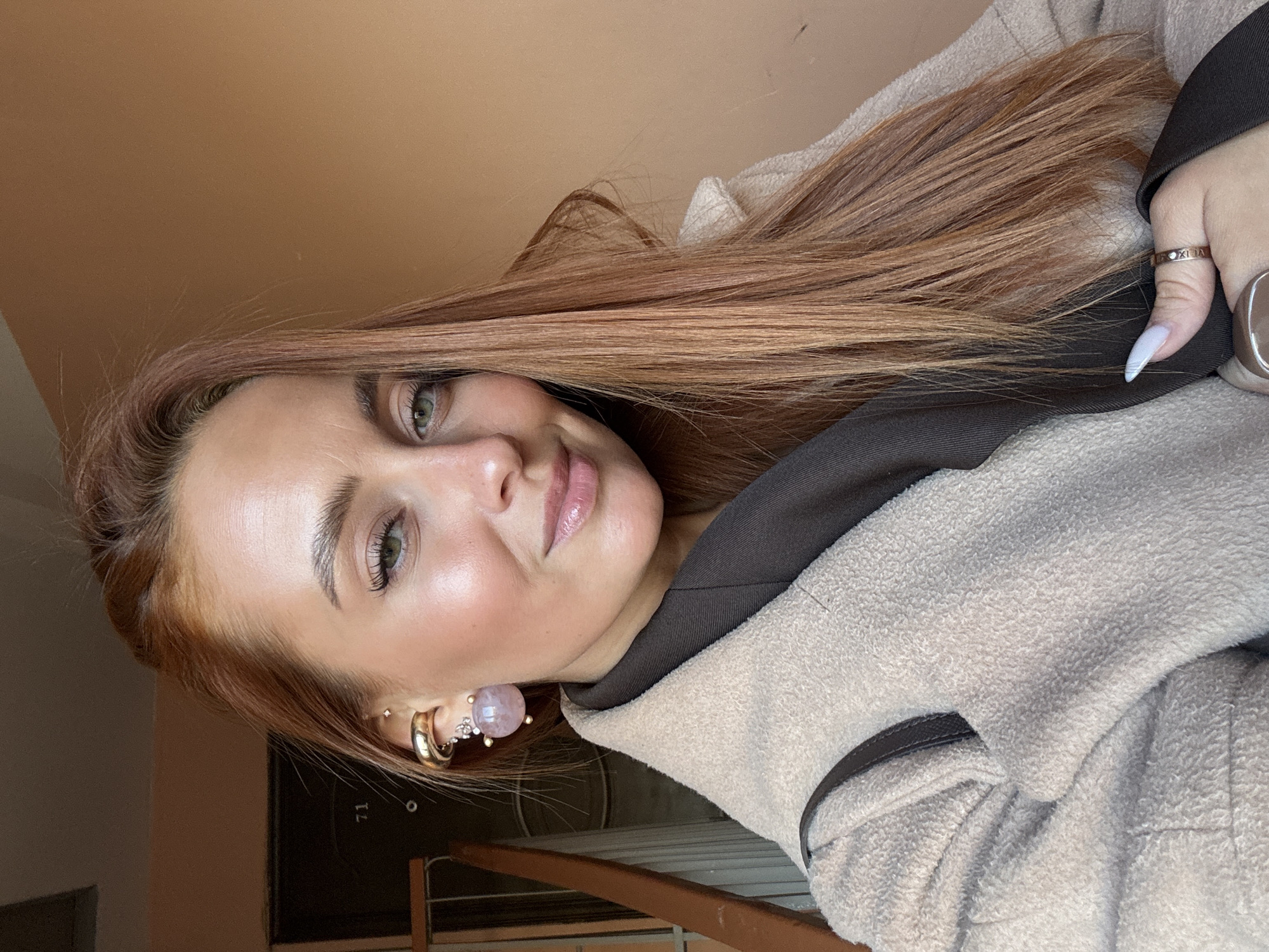 A young woman with long, straight, reddish-brown hair and green eyes poses for a selfie, looking directly at the camera with a soft smile. Her skin has a dewy finish, and she wears light makeup, including lip gloss and mascara.
She is dressed in a cozy, light beige or cream-colored fleece jacket with a dark brown collar or hood lining. Her left ear is adorned with multiple earrings: a prominent light pink, possibly spherical, stud earring with small sparkly accents, and at least one gold-toned hoop earring. On her right hand, a ring with visible Roman numerals, possibly "XLIX," is worn on her index finger, along with a larger, darker ring on her middle finger. Her nails are long, pointed, and painted a light, possibly white or pale blue, color.
The setting appears to be indoors, likely a residential or public building in Chaikovsky, Russia, during daytime, indicated by the natural light. The background features a plain, light brown/tan wall. A dark surface with the number "71" faintly visible can be seen behind her. In the lower part of the image, a wooden or metal railing, possibly part of a staircase or balcony, is visible, suggesting a hallway or entrance area. The overall mood is calm and pleasant.