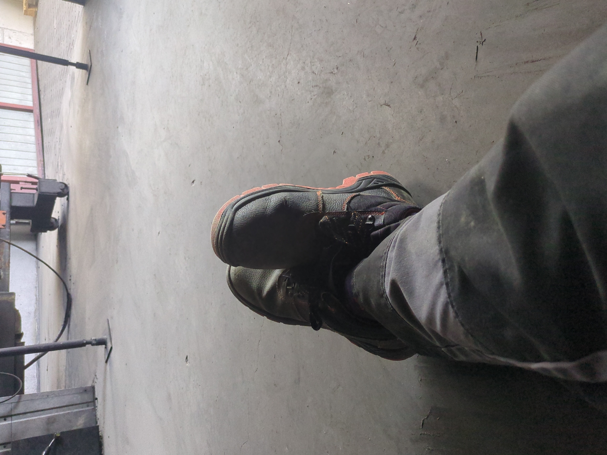 The image shows a close-up view of a person's legs and feet wearing work pants and safety boots, resting on a concrete floor. The scene appears to be indoors, possibly in a workshop or industrial setting, given the presence of heavy machinery and a grey, utilitarian environment. The concrete floor is scuffed and shows some signs of dirt and wear. In the background, parts of machinery, including metal legs and possibly a forklift, are visible. The lighting is dim, suggesting an overcast day or an indoor space with limited natural light. The specific location in Bolesławów, Poland, is not discernible from the visual information provided in the image alone.
