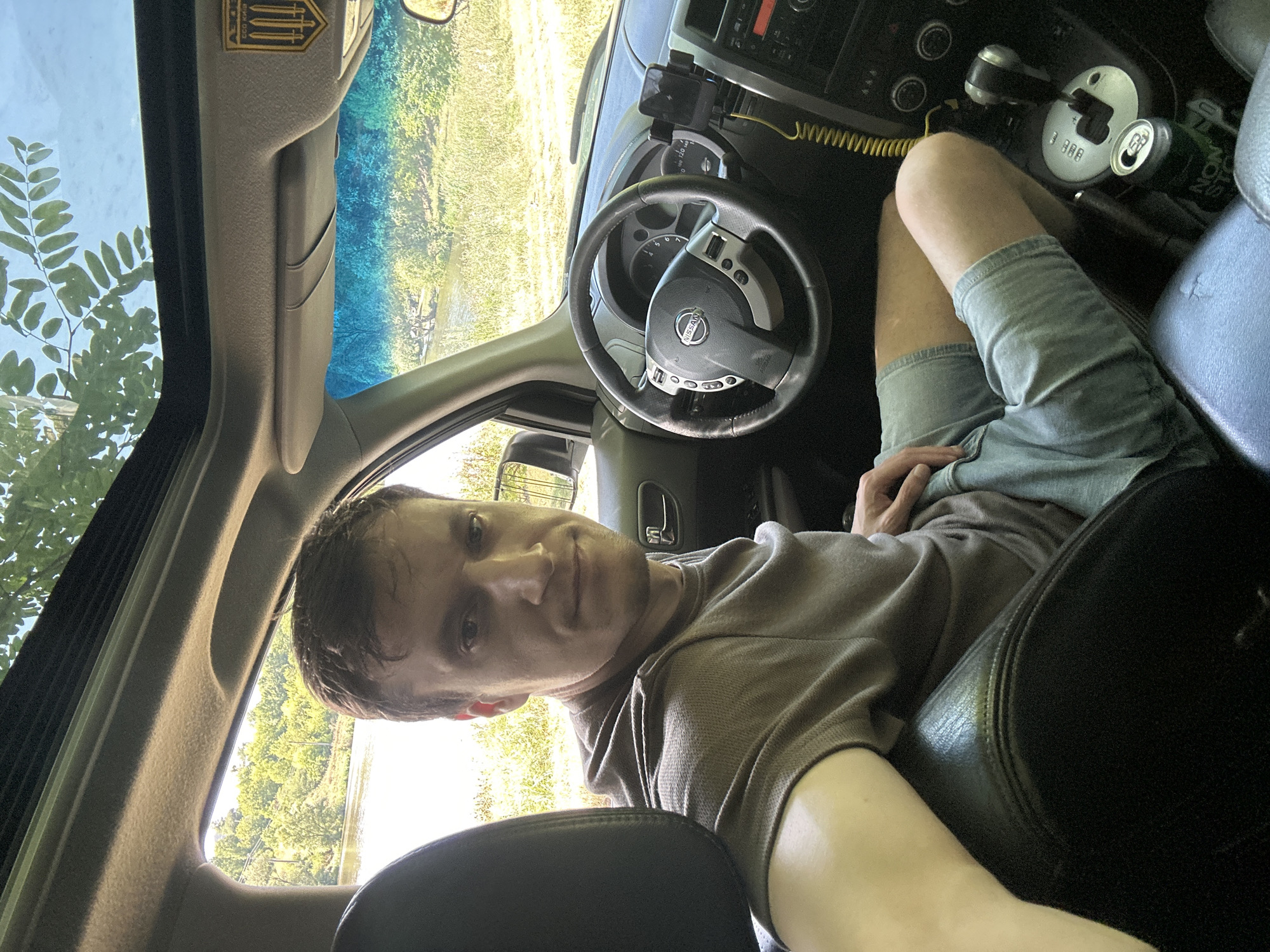 A man with short brown hair is sitting in the driver's seat of a car. He is wearing a brown t-shirt and light blue shorts. The car's interior is visible, including the steering wheel, dashboard, and gear shift. A smartphone is mounted on the dashboard. Outside the car, there is a body of water and trees. The scene appears to be located in Makiv, Ukraine, as indicated by a badge on the sun visor that features the Ukrainian trident.