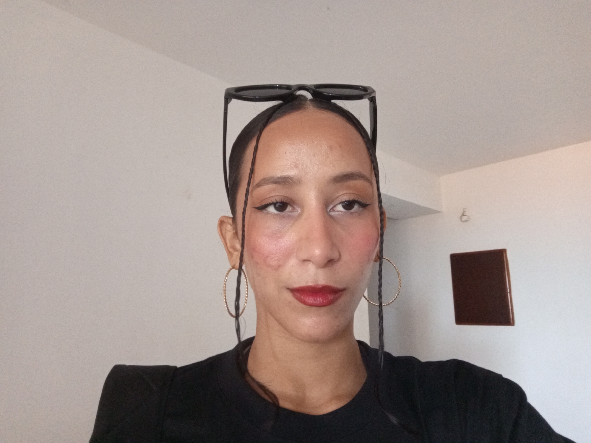 A person with brown skin, wearing a black shirt, gold hoop earrings, and sunglasses on top of their head, is centered in the frame. The person has dark hair with two braids on each side of their face and is wearing makeup, including eyeliner and red lipstick. The background is a plain white wall with a small brown square object hanging on the right side. The location is not discernible as Porlamar, Venezuela. There are no discernible actions or interactions.