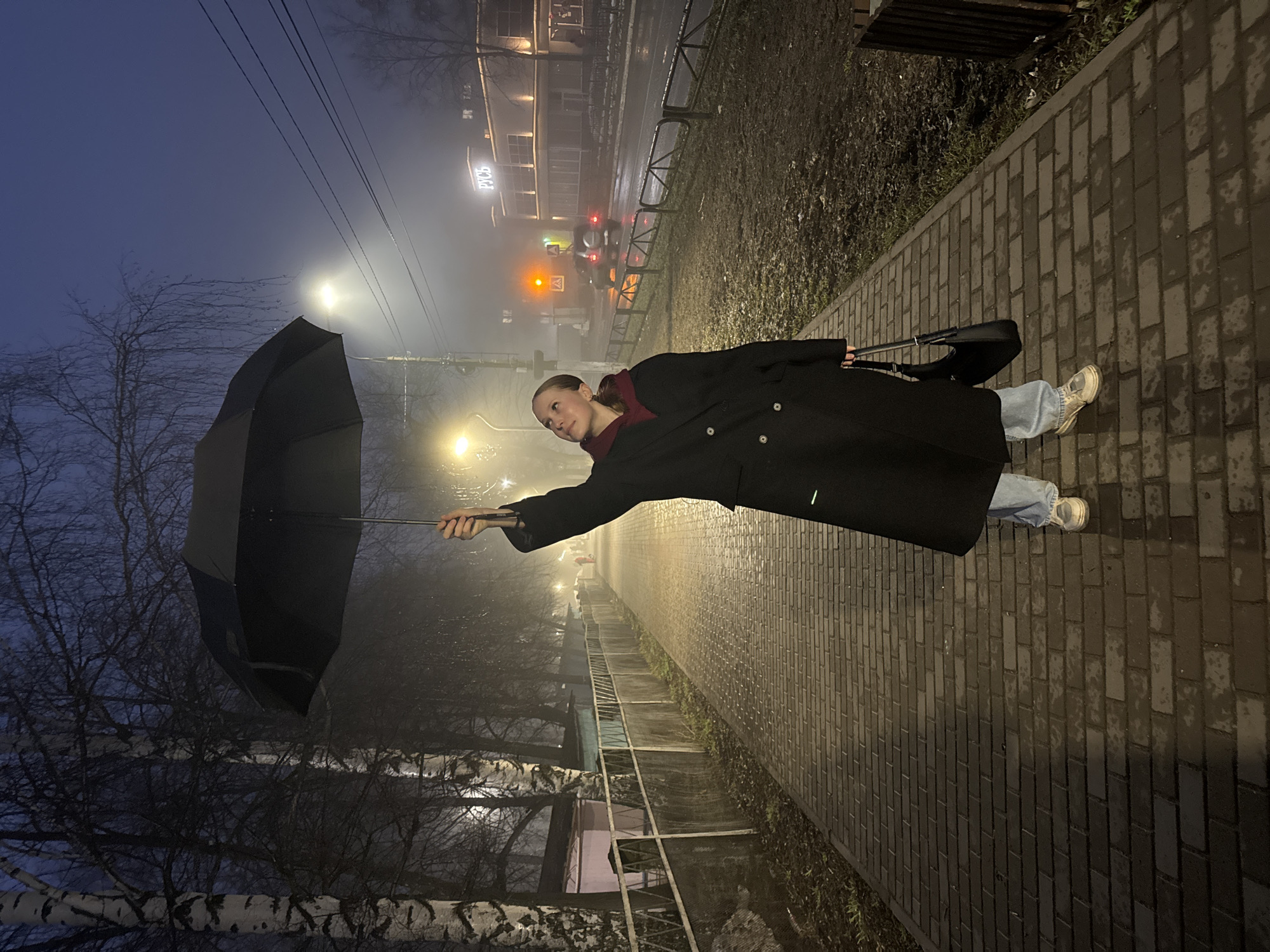 A woman in a dark coat and light blue jeans stands on a brick sidewalk in Chaikovsky, Russia, holding a black umbrella. The sky is foggy and dim, and streetlights illuminate the scene. Buildings are visible in the background. The woman is looking toward the light with her body angled toward the right of the frame. A black purse hangs from her shoulder.