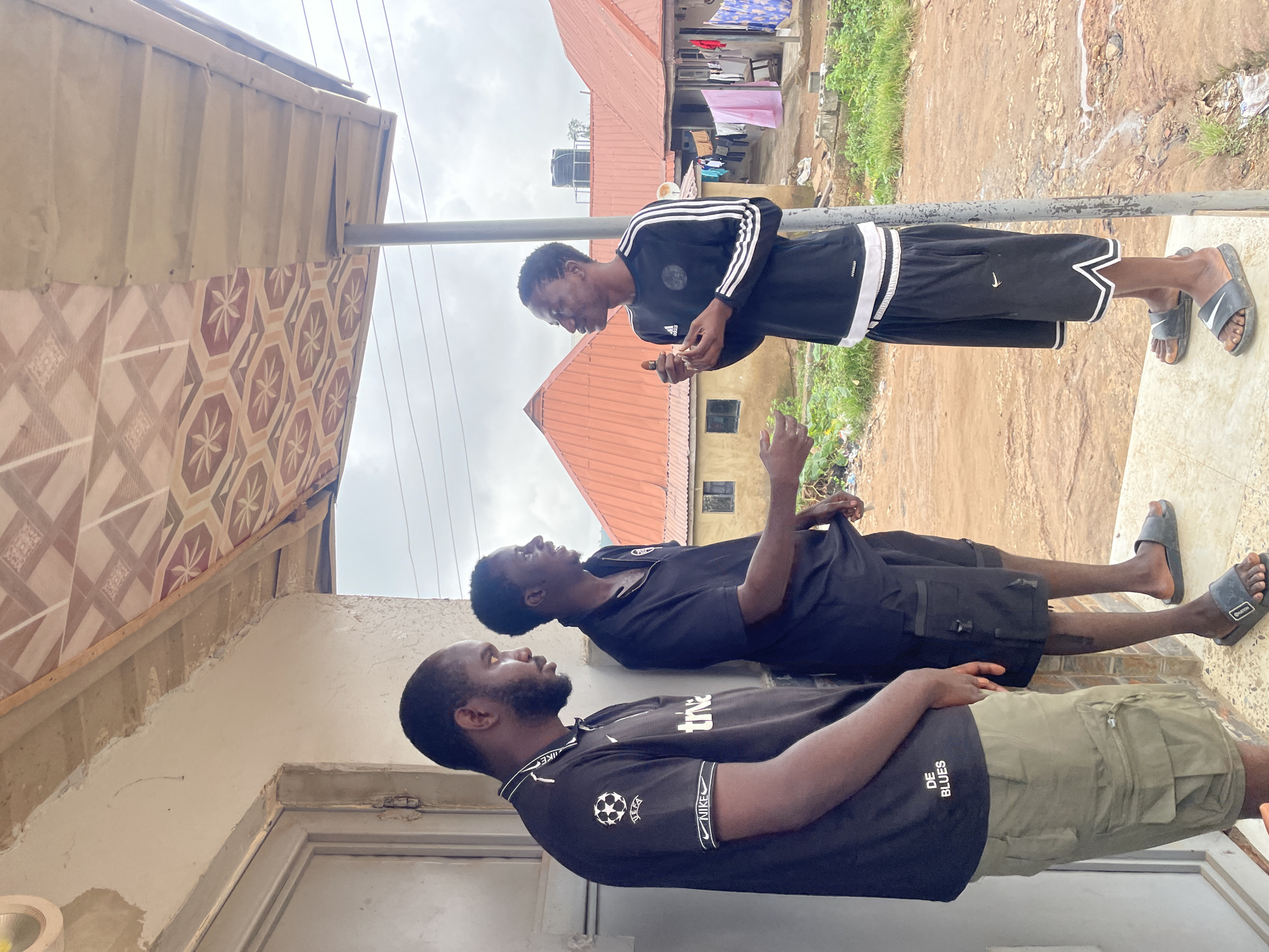 Three men are standing on a raised concrete platform outside a building with a corrugated metal roof adorned with patterned tiles. The man on the left is wearing a black t-shirt with white trim and a soccer ball logo, olive green cargo shorts, and is looking down and to his right. The middle man, in a black t-shirt and black shorts, is facing the man on the right and gesturing with his hands as if in conversation. The man on the right, wearing a long-sleeved black shirt with white stripes and black shorts with white trim, is holding a small object in his right hand and looking at the middle man. In the background, there are residential buildings with orange corrugated roofs, clothes hanging on a line, and overgrown vegetation. The ground appears to be dirt and mud. The scene is set in Kurunduma, Nigeria.