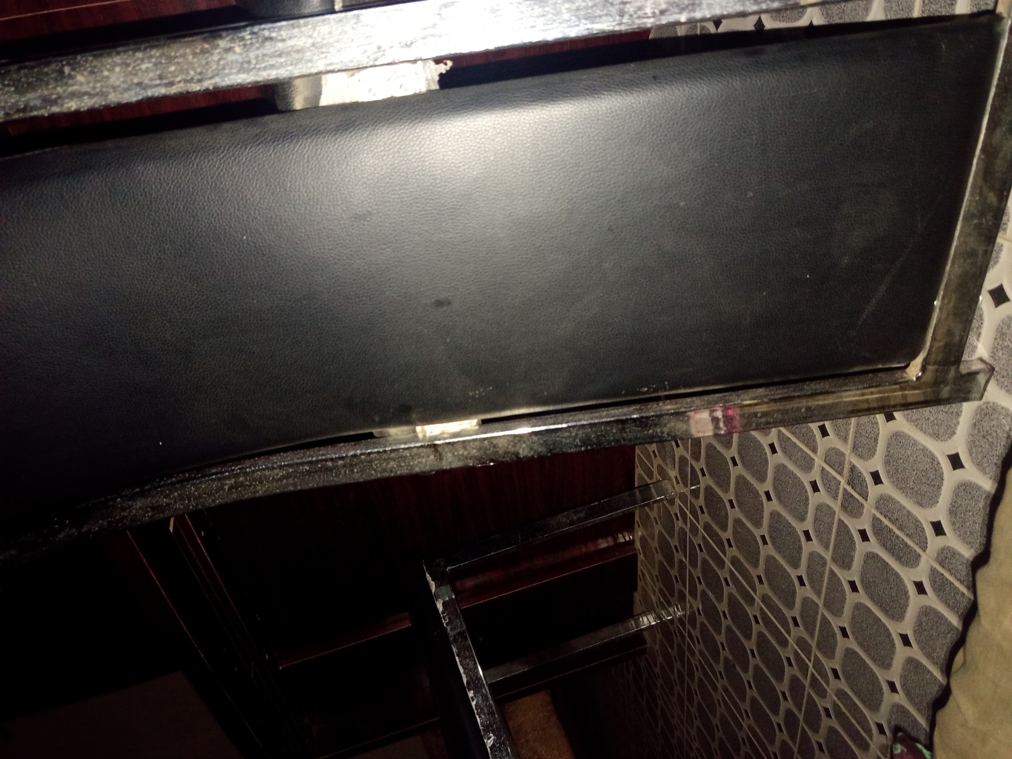 The image shows a close-up, angled view of a black faux leather chair with a metal frame. The chair is situated on a patterned tiled floor. In the background, a dark wooden cabinet or dresser is visible. The scene appears to be indoors, possibly in a residential or commercial setting. The lighting is dim, creating shadows. The location is Jalingo, Nigeria.