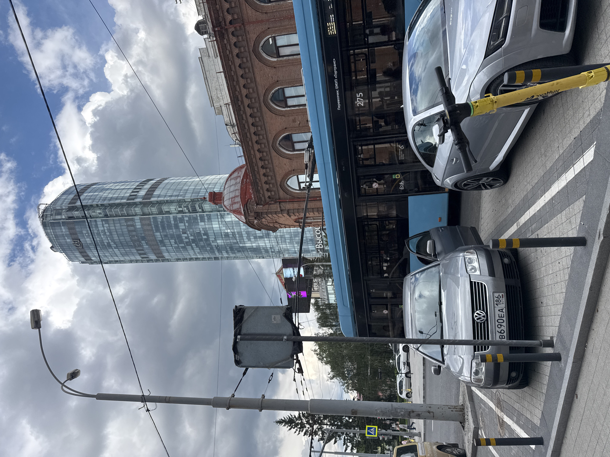An urban scene in Yekaterinburg, Russia, features a modern glass skyscraper dominating the background, with a red-domed historic brick building to its right. A blue trolleybus is parked on the street, with its number "275" visible. In the foreground, a silver Volkswagen sedan is parked in a marked spot, its driver's side door open. To its right, a silver Audi coupe is parked. A yellow electric scooter is positioned in front of the Audi, leaning against a bollard. A traffic light and a street lamp are also visible. The sky is partly cloudy.