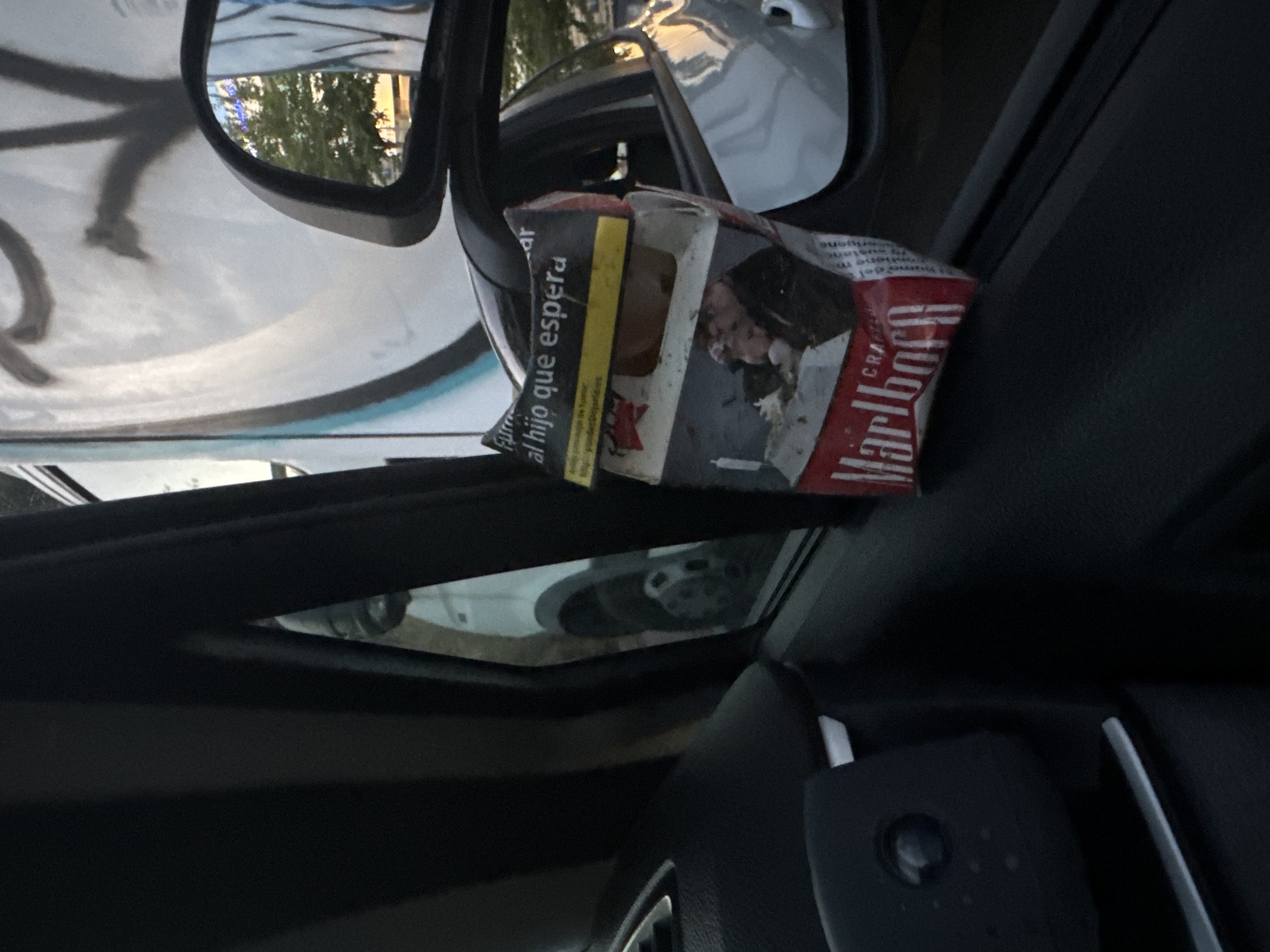 A crumpled Marlboro cigarette pack is visible inside a car, resting on the door panel near the side mirror. The pack displays text in Spanish, including a warning about smoking during pregnancy. In the reflection of the side mirror, a building with graffiti and trees can be seen. The interior of the car appears to be dark, with parts of the steering wheel and dashboard visible.