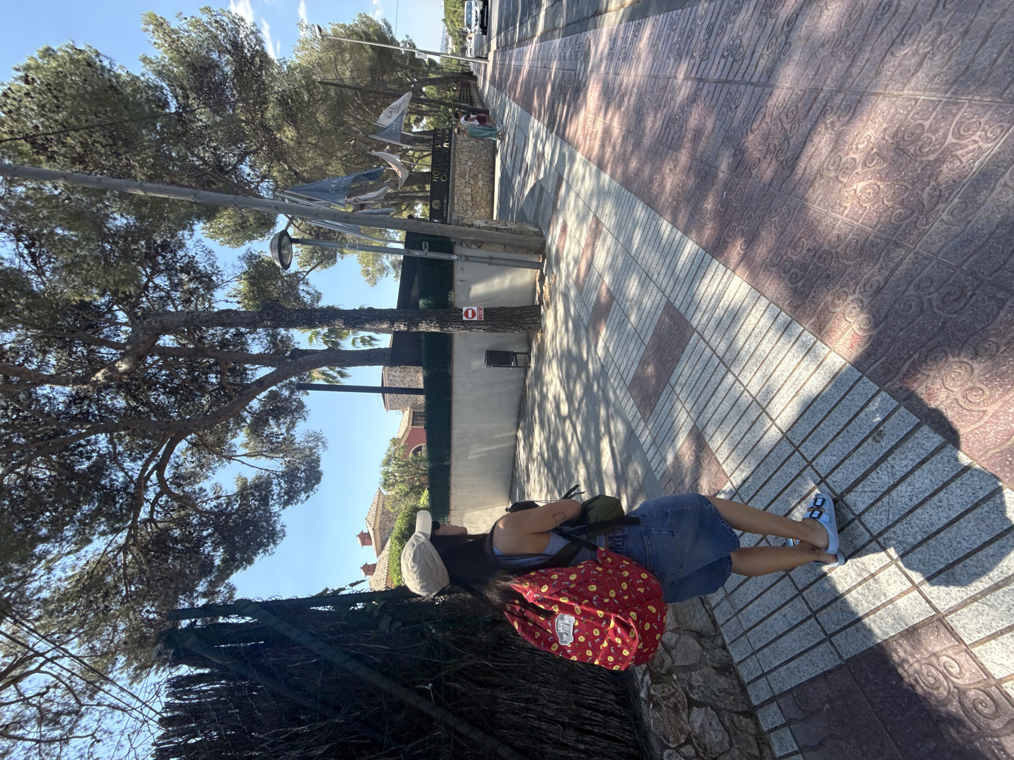 A young woman wearing a beige baseball cap, a light blue tank top, and denim shorts stands on a tiled sidewalk next to a street in Platja d'Aro, Spain. She has a red backpack with a yellow pattern and a small green bag slung over her shoulder. She is looking to her right, away from the camera. In the background, a street lined with trees runs alongside the sidewalk. To the left of the sidewalk is a stone wall and a dark fence. Across the street, a hotel with flags and palm trees is visible. A white car is driving down the street in the distance. The sun casts shadows on the sidewalk, indicating it is daytime.