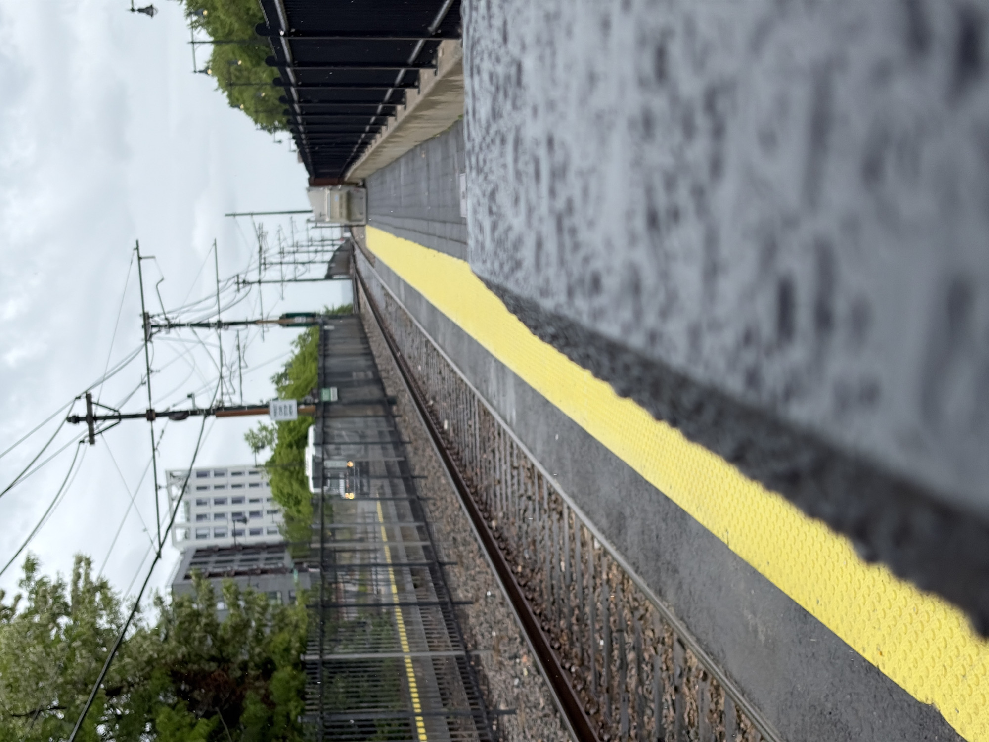 The image depicts a train station in Boston, Massachusetts, with the view along the tracks. The platform is wet, indicating recent rain. A yellow safety line marks the edge of the platform. Electrical wires and support structures are visible overhead, characteristic of a train line. In the background, buildings and trees are visible. The photo is taken at an angle from near the front of the platform which gives a strong depth of field.