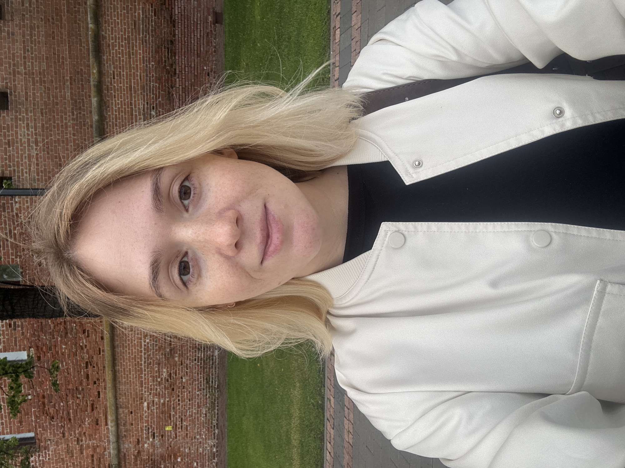 A young woman with blonde hair and freckles smiles directly at the camera. She is wearing a white bomber jacket over a black shirt. She has a brown strap from a bag visible over her right shoulder. In the background, there is a red brick wall and a patch of green grass. The ground in front of her appears to be paved with bricks. The scene suggests an outdoor setting in the city of Nizhny Novgorod, Russia, possibly near a historical building or fortification due to the brick architecture.