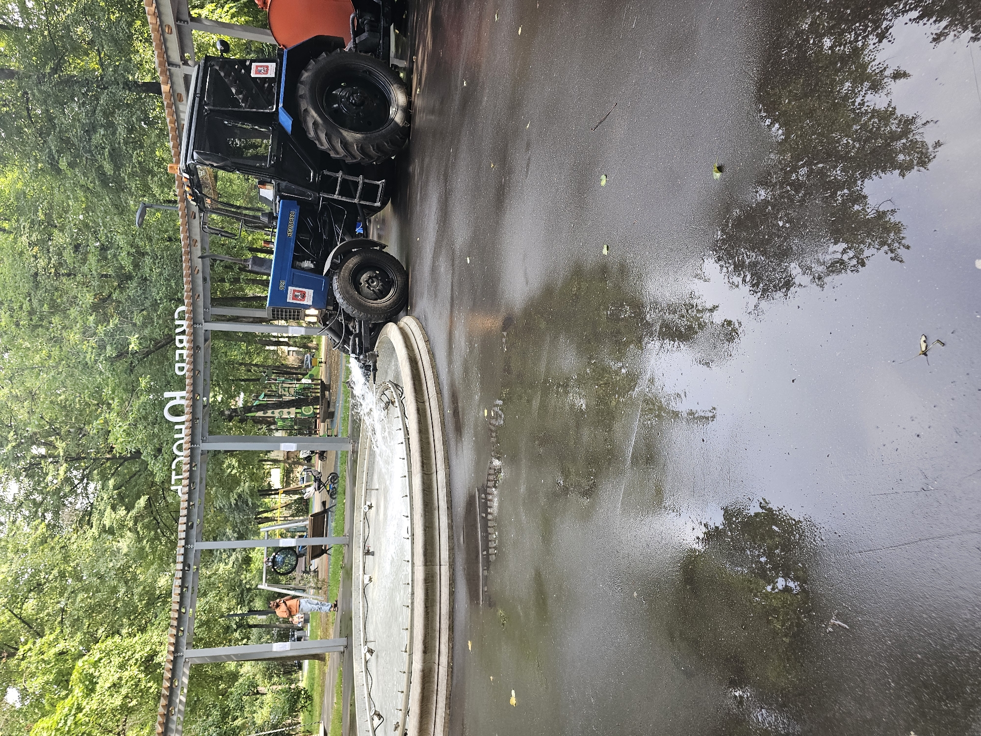 In a park setting, a blue and black tractor is positioned near a circular fountain, from which water is being sprayed. The tractor has a visible label reading "BELARUS" on its side, and it appears to be connected to an orange water tank. The ground around the fountain is wet, with reflections of the surrounding trees and structures visible on the surface. In the background, a man in an orange shirt stands near a park bench and playground equipment. Above, a canopy structure with wooden beams is visible. The environment suggests an outdoor public space, likely a park or recreational area in Khimki, Russia, given the presence of a Belarus tractor, commonly used in the region.