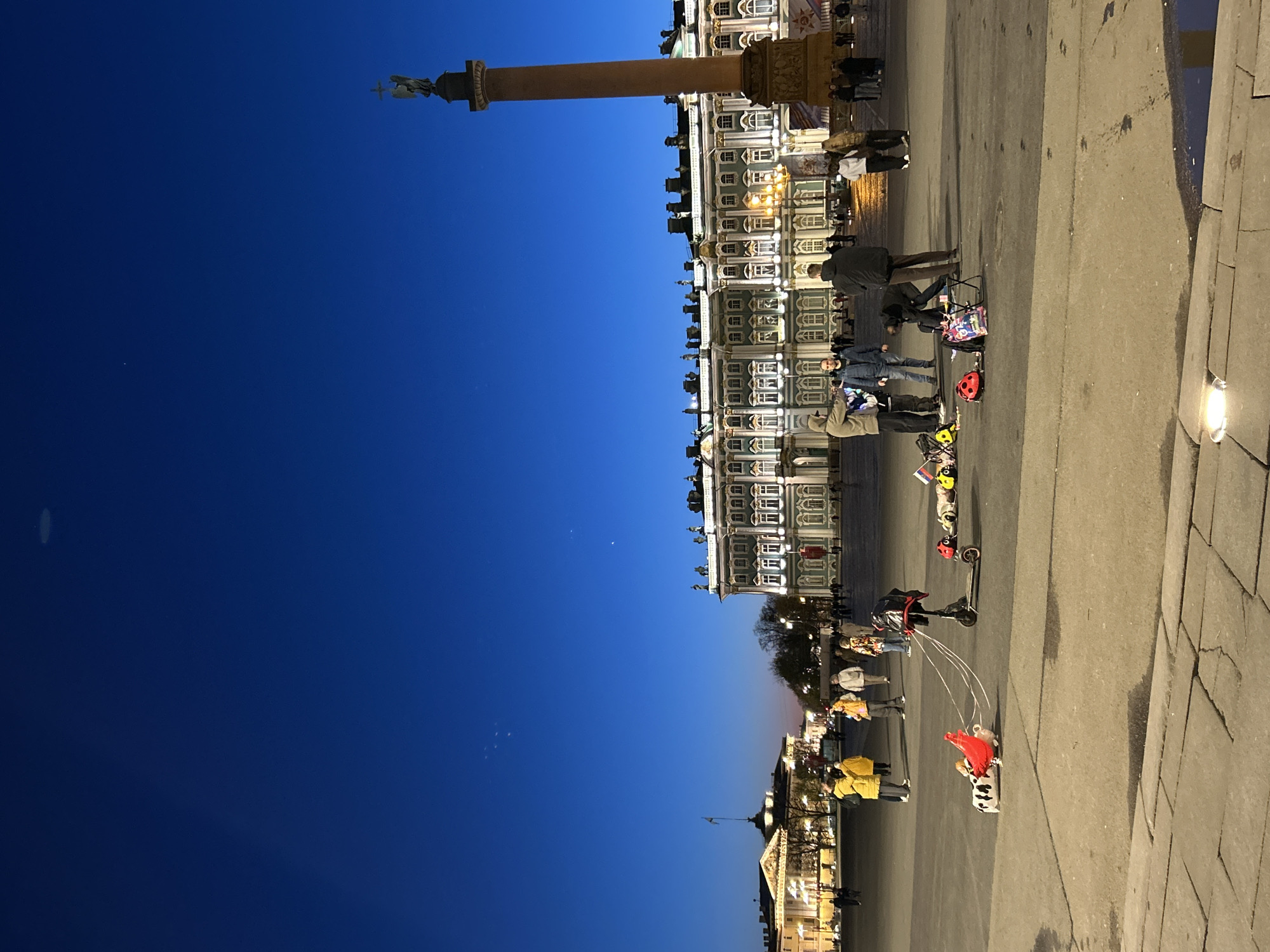 The image depicts Palace Square in Saint Petersburg, Russia, at dusk. The Alexander Column stands tall on the left of the frame, against a deep blue sky. The Winter Palace is positioned to the right. Pedestrians are seen strolling across the square. Several individuals are grouped together, one kneeling near a red ladybug helmet, next to small scooters. Streetlights are visible along the edge of the square. The ground is paved with large stone blocks.