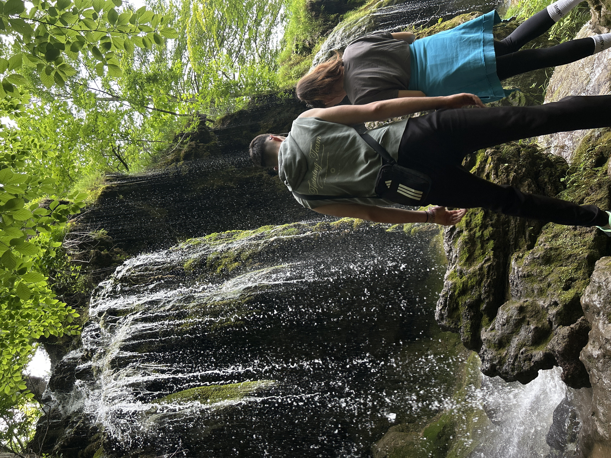 This image captures a natural outdoor scene featuring a waterfall cascading over a moss-covered rock face, located in the city of Unknown, Unknown. Two individuals are present, viewed from behind, standing near the base of the waterfall.
The individual on the left, appearing male, wears a sleeveless olive-green hooded sweatshirt with the text "Training Team" printed in white on the back. He also wears dark-colored trousers and a black Adidas shoulder bag with white stripes positioned across his chest and left hip. His left arm is slightly extended. The individual on the right, appearing female with reddish-brown hair, wears a grey t-shirt, a light blue wrap-around skirt or cover-up over black leggings, and white socks. Both individuals face the waterfall, observing the cascading water.
The waterfall is a prominent feature, with water actively falling over a dark, irregularly shaped rock face heavily covered in vibrant green moss. Water spray is visible at the base of the falls. Lush green foliage and tree branches are visible above and to the right of the waterfall, suggesting a dense forest or heavily vegetated area. The ground around the base of the waterfall is rocky and appears wet, also adorned with moss. The lighting is diffused, indicating an overcast day or light filtering through a dense canopy.