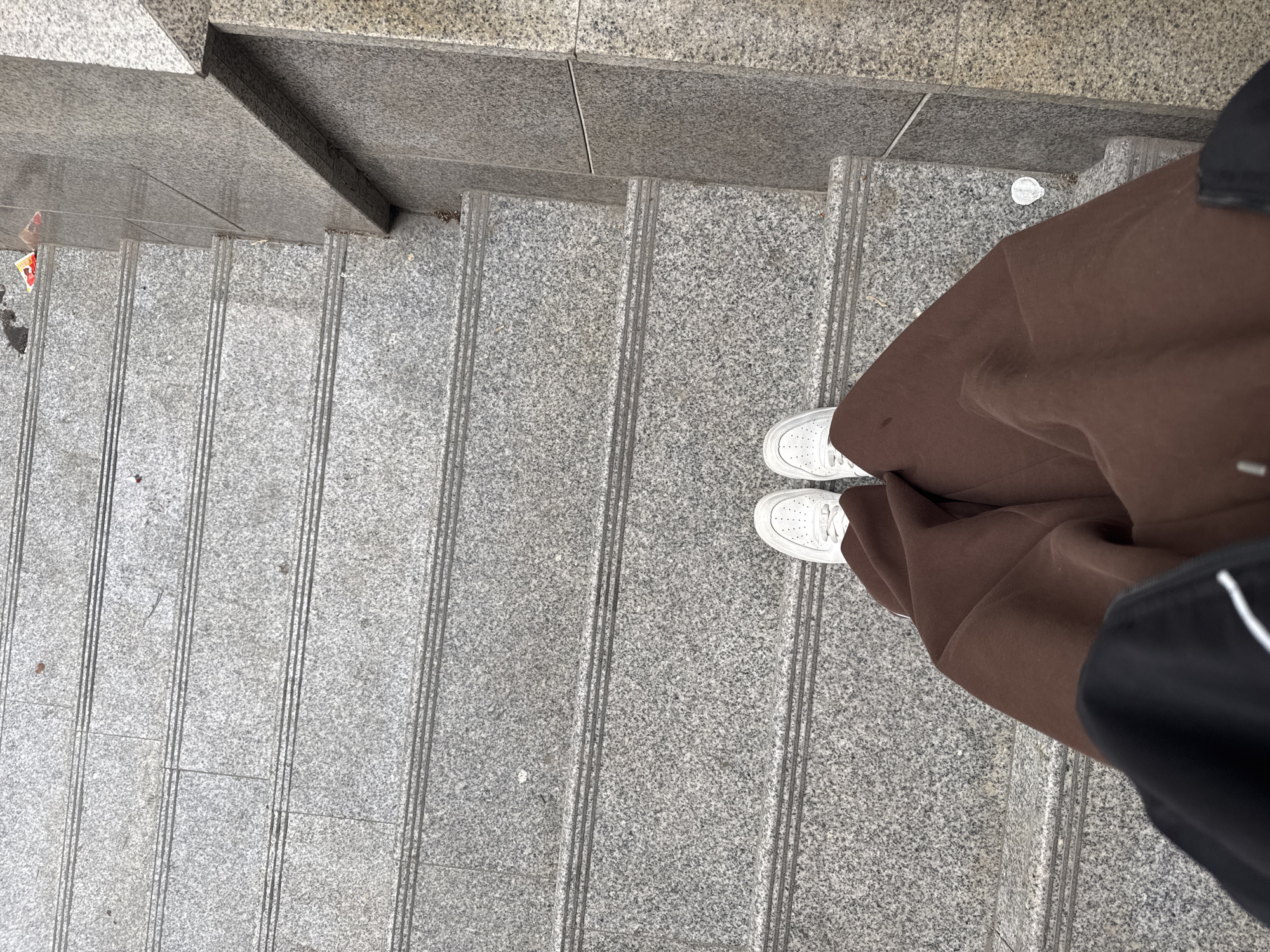 The image shows a view from above looking down onto a staircase. The staircase is made of grey, speckled stone or concrete. A person is standing on the steps, wearing white sneakers and a brown garment that appears to be a long skirt or dress. The lower right of the image also shows a partial view of a black bag or clothing item with a white stripe.
The location in Azov, Russia, is unknown.
