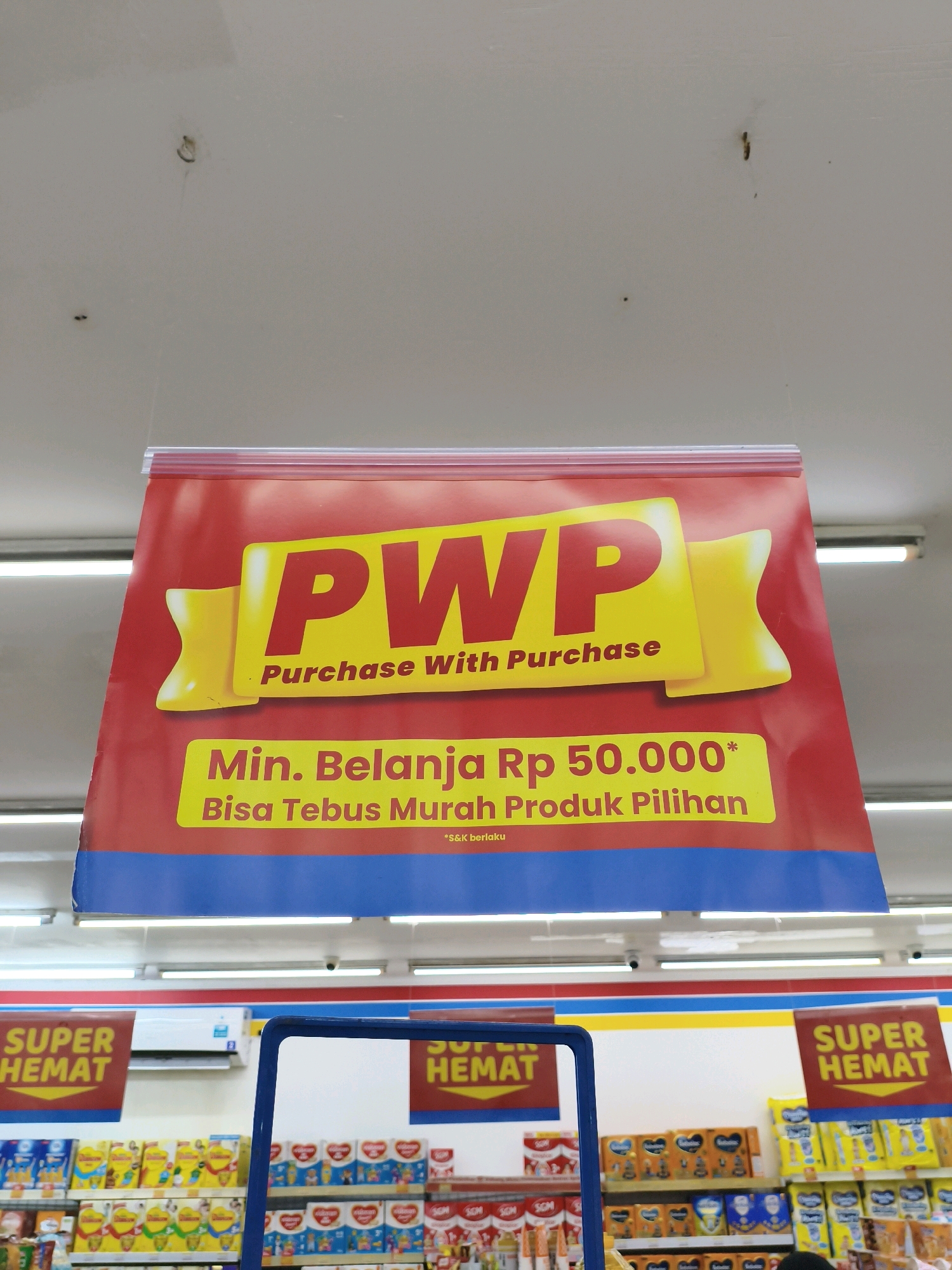 A red banner hangs from the ceiling in a retail store, advertising a "Purchase With Purchase" (PWP) promotion. The banner prominently displays "PWP" in large yellow letters with a yellow ribbon graphic. Below this, text in Indonesian reads "Min. Belanja Rp 50.000" (Minimum spending Rp 50,000) and "Bisa Tebus Murah Produk Pilihan" (Can redeem cheap selected products). A smaller note at the bottom says "*S&K berlaku" (*T&C apply). In the background, shelves stocked with various products are visible, along with signs that say "SUPER HEMAT" (SUPER SAVINGS). An air conditioning unit is mounted on the wall. The lighting suggests an indoor commercial environment. The location is identified as Balesono, Indonesia.