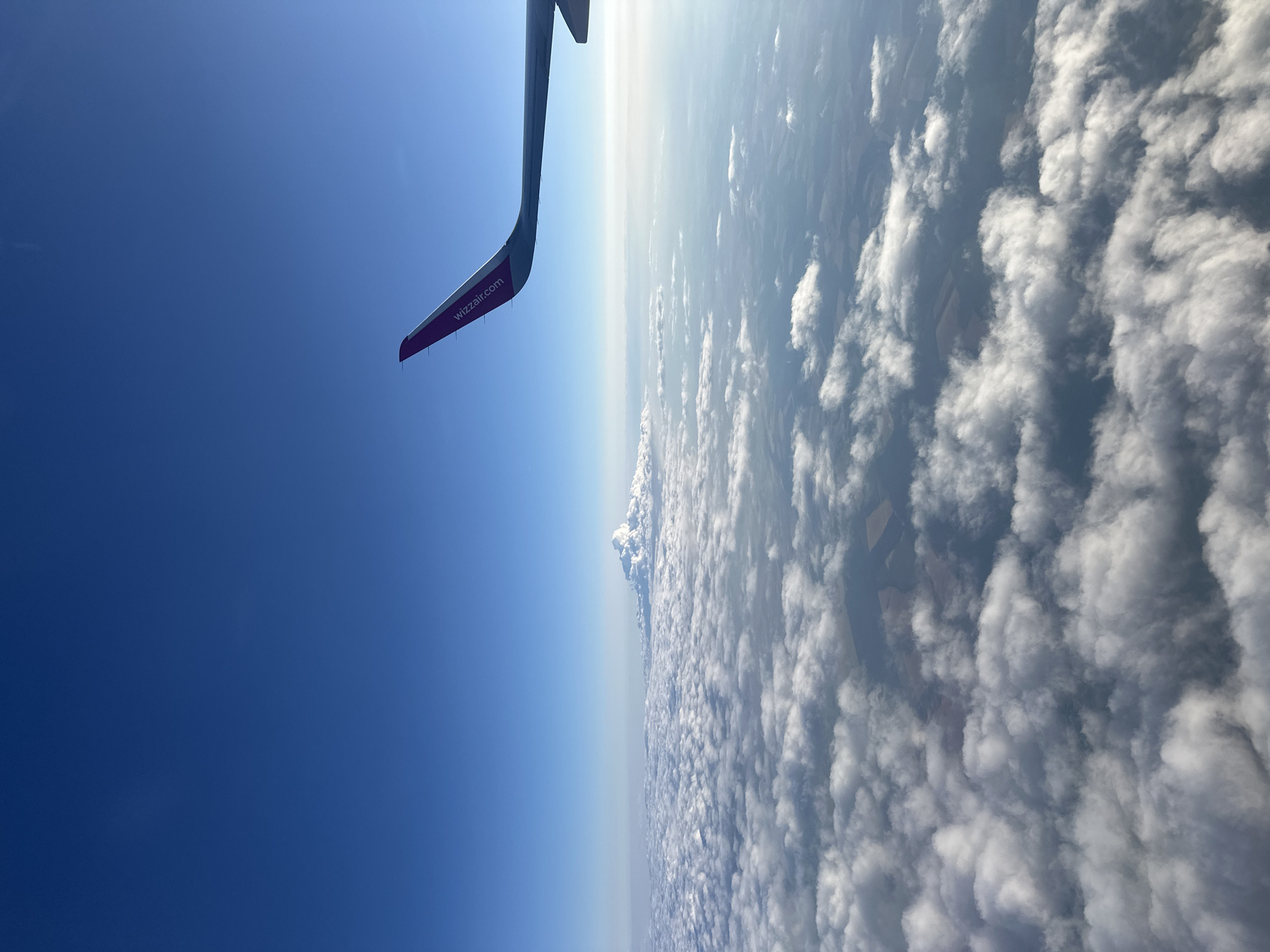 An aerial view from an airplane window shows a vast expanse of white clouds below a clear blue sky. The wing of the airplane, featuring the "wizzair.com" logo in purple, is visible in the upper right corner. The landscape beneath the clouds is partially visible in some areas, revealing fields and structures, suggesting an overflight of a populated region. Given the presence of Wizz Air and the general landscape, it is plausible that this image was taken during a flight to or from Budapest, Hungary, as Wizz Air is a prominent airline serving that city. There are no people, specific buildings, or identifiable landmarks visible in the image to pinpoint an exact location within Budapest. The environment is predominantly atmospheric, characterized by clouds and sky, with a glimpse of the Earth's surface below.