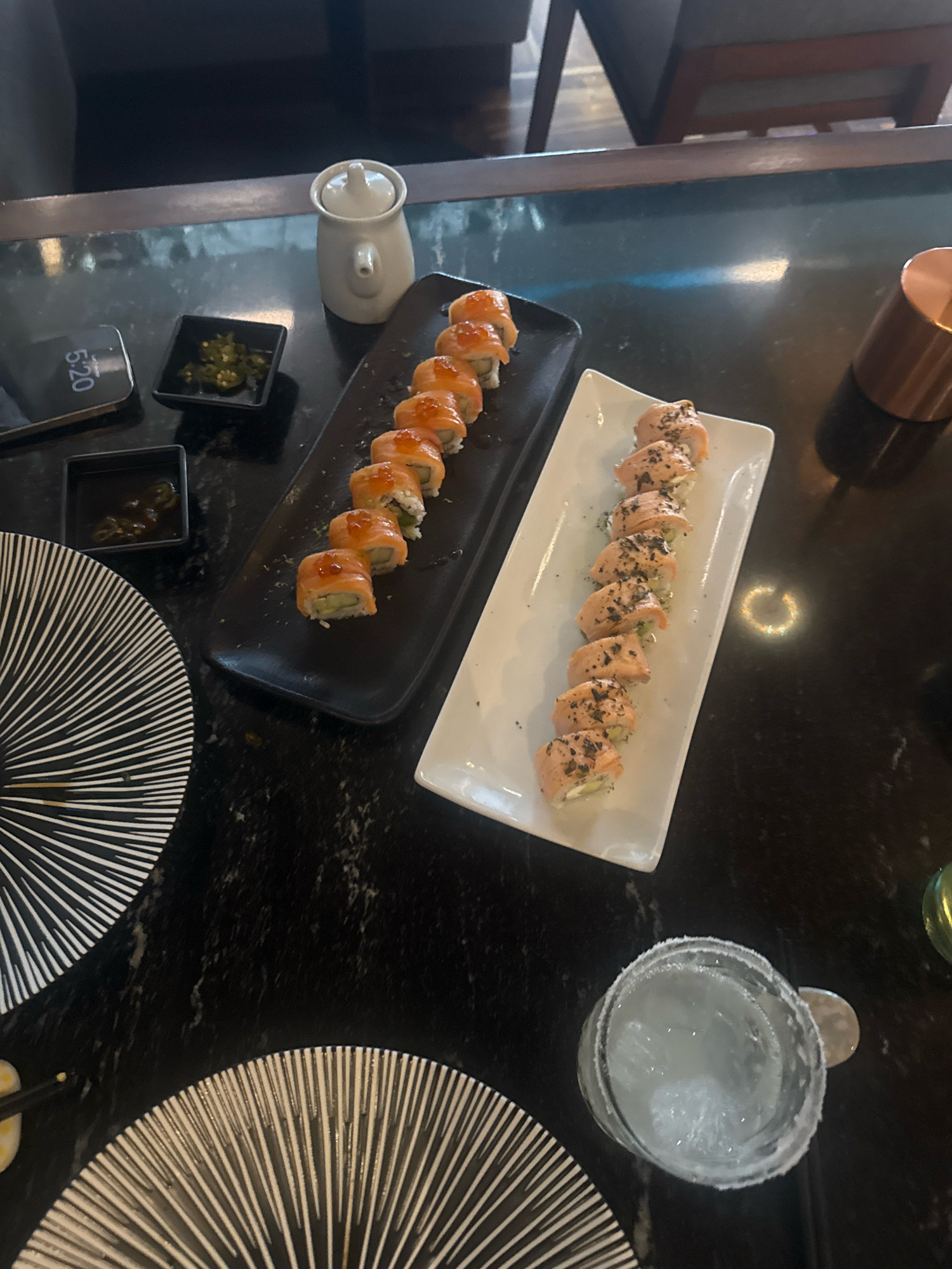The image shows a dark, granite-like table with two plates of sushi rolls, small condiment dishes, and a glass of iced beverage. One plate, a black rectangular dish, contains several sushi rolls topped with roe. Adjacent to it is a small black dish with chopped green peppers and another with a dark sauce. A white ceramic condiment dispenser is also present. The second plate, a white rectangular dish, holds a row of sushi rolls topped with what appears to be seared salmon and black seasoning. A glass with a salt-rimmed edge, filled with ice and a clear liquid, sits to the right of the white plate. Two decorative, fan-shaped placemats with black and white stripes are visible at the bottom of the image. In the background, dimly lit chairs and a table are visible, suggesting an indoor dining setting. The phone screen shows the time as 5:20. The context of Puebla, Mexico, indicates this is likely a restaurant in that city.