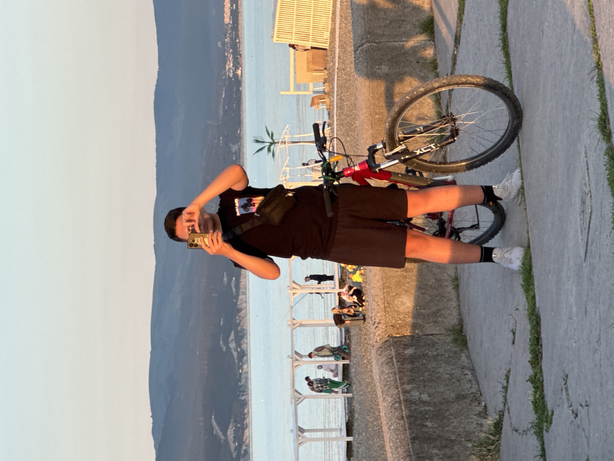 A young person, dressed in a black t-shirt, black shorts, white sneakers, and black socks, is taking a selfie with a smartphone in a bright, outdoor setting. They have a black fanny pack or shoulder bag strapped across their chest. A red and black mountain bike, with "XCT" visible on its front fork, is positioned beside them.
The individual is standing on a concrete promenade or embankment. In the mid-ground, a large body of water, consistent with the Black Sea at Novorossiysk, Russia, stretches towards the horizon. Beyond the water, a range of mountains or hills can be seen under a light, hazy sky. Along the waterfront, several white wooden or metal structures, possibly pergolas, fences, or seating areas, are visible. Various other people are scattered throughout the background, some sitting, some standing, and one person appears to be pushing a small cart or stroller. A small palm tree also dots the landscape.
The photograph captures the scene during the 'golden hour,' likely late afternoon or early evening, characterized by warm, soft light and long shadows stretching across the concrete path. The weather is clear and calm, suggesting a pleasant day for outdoor activities. The overall mood is relaxed and casual, depicting a leisurely moment by the sea.