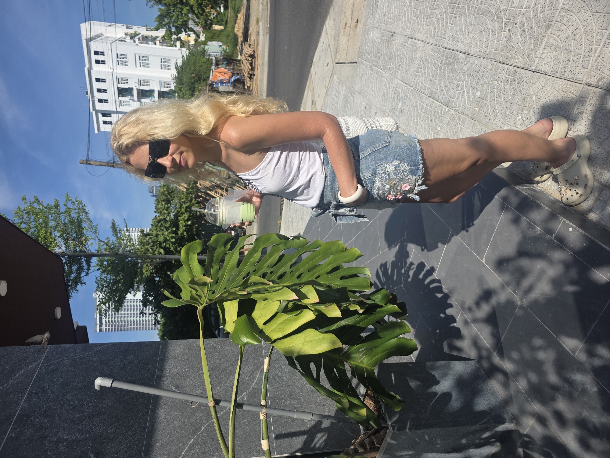 A long-haired blonde woman stands on a sidewalk in what appears to be Đà Nẵng, Vietnam. The woman is wearing a white tank top, a denim skirt, and decorative Crocs. She is also wearing sunglasses and a white bracelet and holding a green drink. The sidewalk is made of patterned grey tiles, and a potted Monstera plant is next to it. Behind the woman is a street and a multi-story white building with rectangular windows and a blue sky. Other buildings and trees are visible in the distance. The overall setting appears to be an urban, outdoor environment.