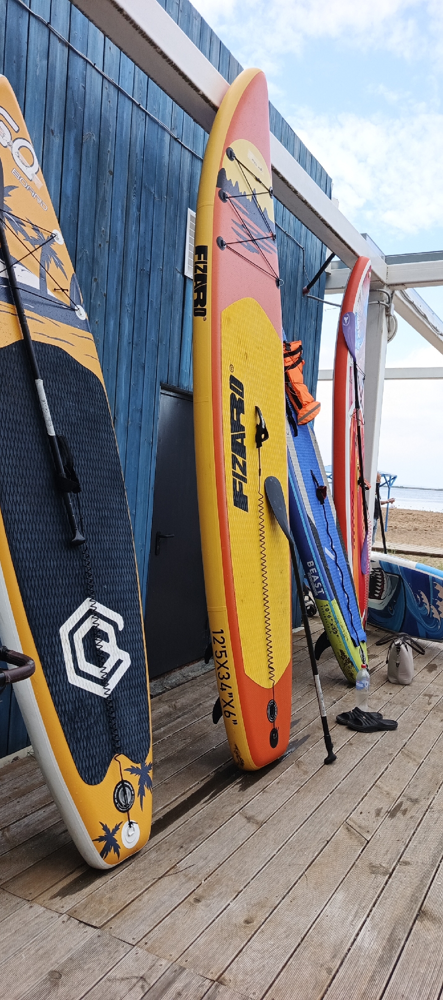 This is a photograph of several stand-up paddleboards (SUPs) leaning against a blue wooden wall, likely at a recreational area or rental shop. The SUPs are colorful, with designs featuring palm trees, geometric patterns, and text. One paddleboard, predominantly yellow and orange, has the brand name "FIZARO" printed vertically. Another paddleboard, with a dark blue deck and yellow trim, displays a white hexagonal logo. A life vest in orange is placed on one of the SUPs.
The SUPs are positioned on a wooden deck, which appears wet in places, suggesting recent use or damp weather. Scattered on the deck are a pair of black flip-flops, a water bottle, and a light-colored bag. In the background, a sandy beach and a body of water are visible, along with a portion of a white structure and the sky with clouds.
While the image shows a setting conducive to water sports, there is no direct evidence linking it specifically to the city of Chaikovsky, Russia. The details provided in the image (e.g., signs, architecture, landscape) do not contain any identifiable Chaikovsky-specific landmarks or text.