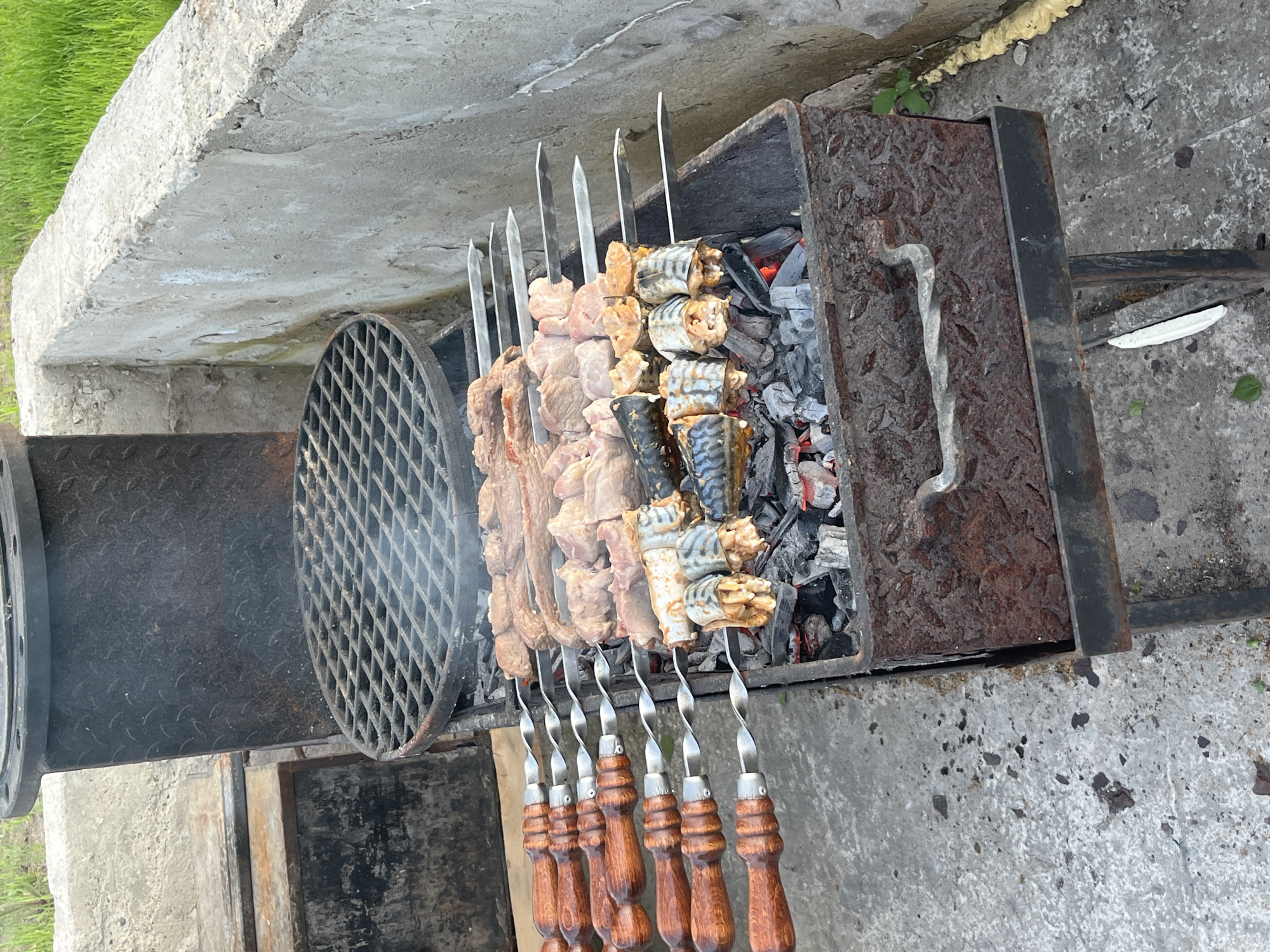 The image shows a barbecue grill in Новая Чернушка, Russia. Skewers of meat and fish are being cooked over hot coals. The grill is made of metal and has a handle on the side. The background is a concrete surface.