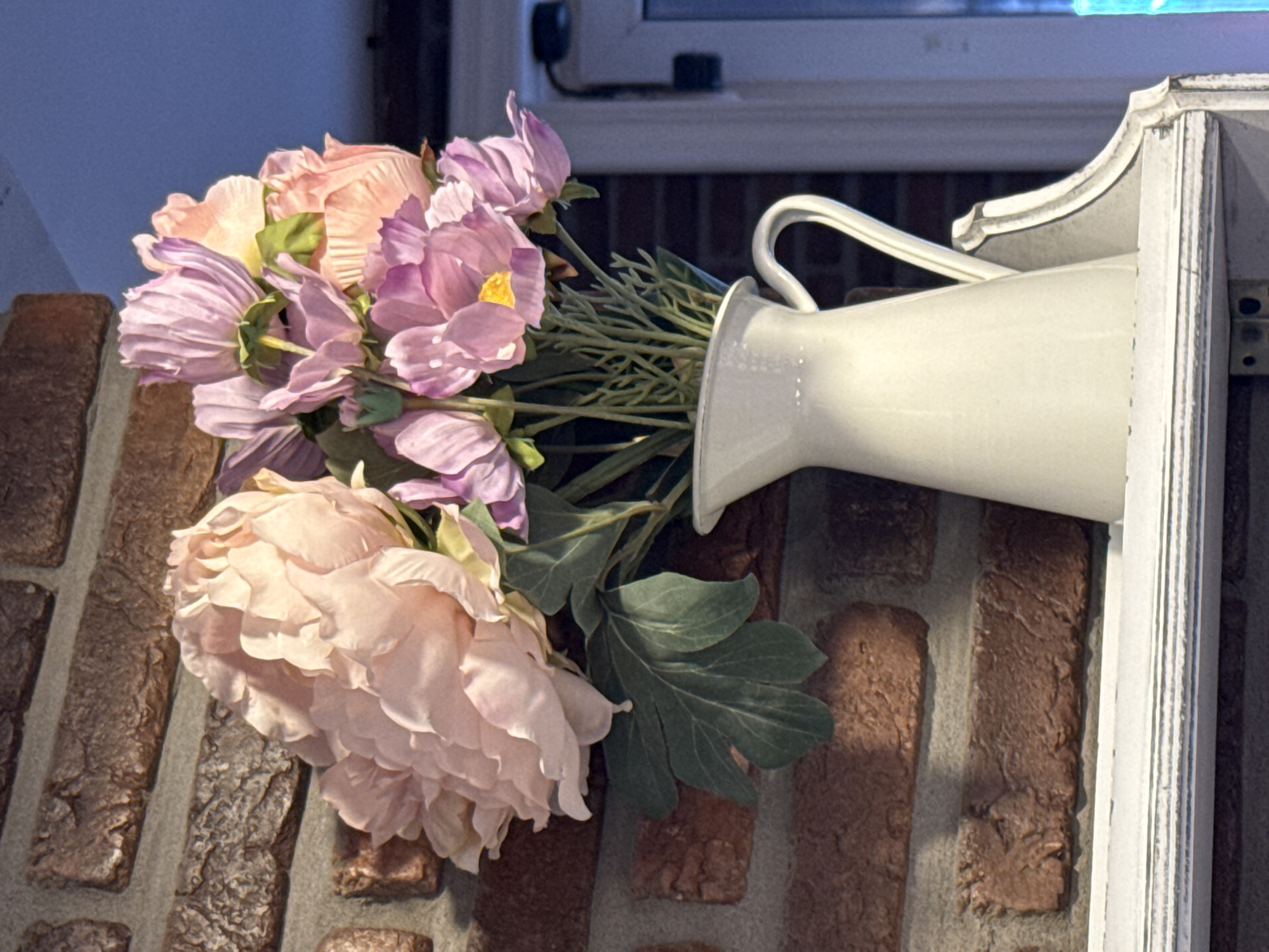 This is a close-up, eye-level, horizontal shot of artificial flowers in a decorative white pitcher. The flowers are a mix of pale pink and lavender peonies and other blooms with green foliage. The pitcher is resting on a brick wall, and a white architectural detail is visible to the right of the pitcher. The background shows a window with blinds. The scene appears to be indoors or in a sheltered outdoor area. The lighting suggests it might be daytime. There is no text visible in the image.