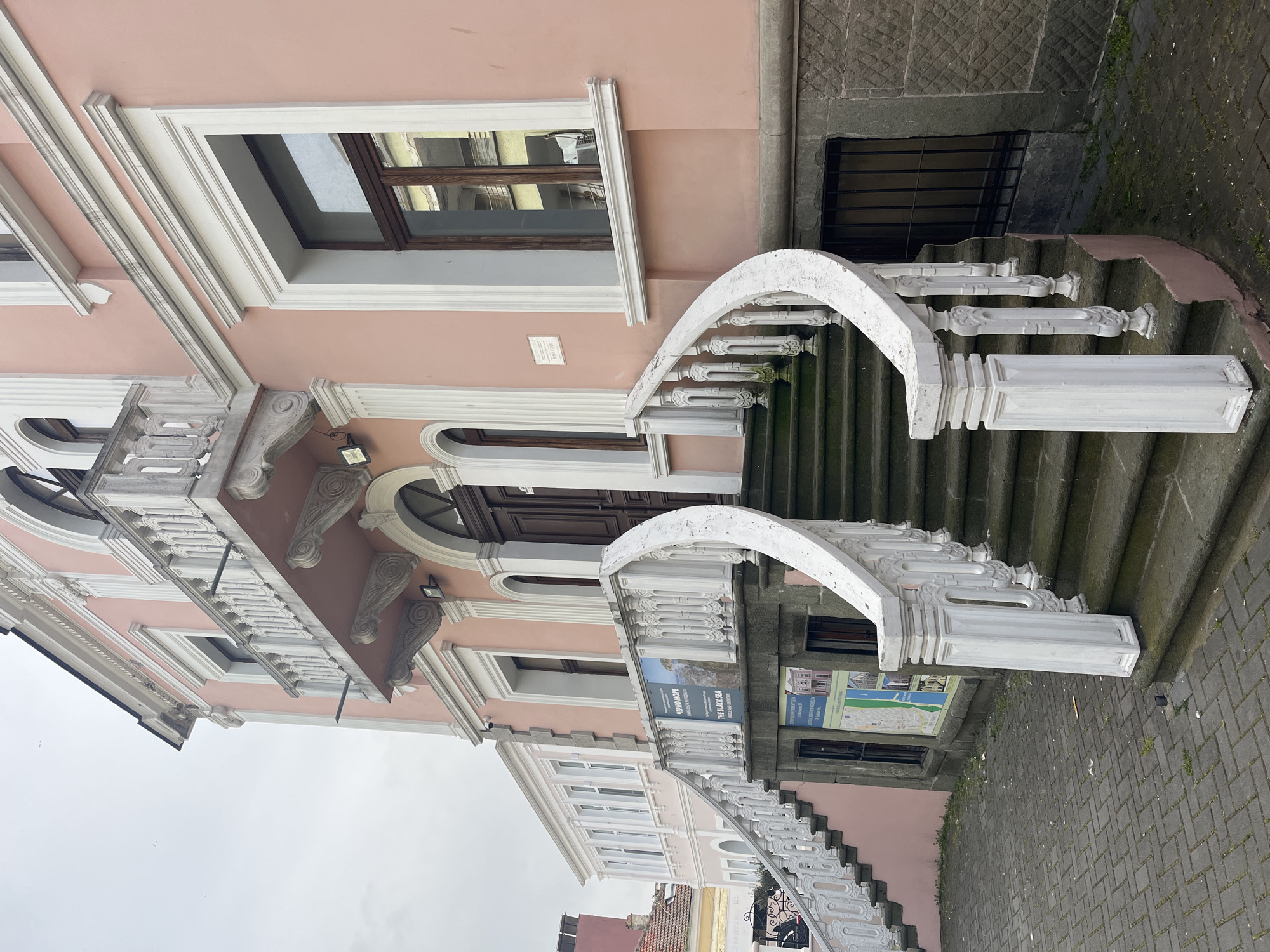 An ornate, light pink building with elaborate white architectural details stands prominently under an overcast sky. The facade features white decorative trim around windows, cornices, and balconies, which are supported by sculpted corbels. A grand, curving staircase made of grey stone steps with decorative white balustrades leads up to the main entrance, characterized by a dark wooden arched double door.
On the left side of the main staircase, near its base, a sign is visible, displaying text in both Bulgarian and English: "ЧЕРНО МОРЕ" and "THE BLACK SEA", along with the word "MUSEUM". A smaller, white rectangular plaque is affixed to the pink wall above the staircase, indicating the building's designation, which appears to be part of the "REGIONAL HISTORICAL MUSEUM BURGAS".
The ground directly in front of the stairs is paved with grey rectangular bricks. Parts of the stone steps and balustrades show signs of weathering, including some moss and grime. The overall scene depicts a historic or significant public building during daytime, with no individuals or specific activities visible.