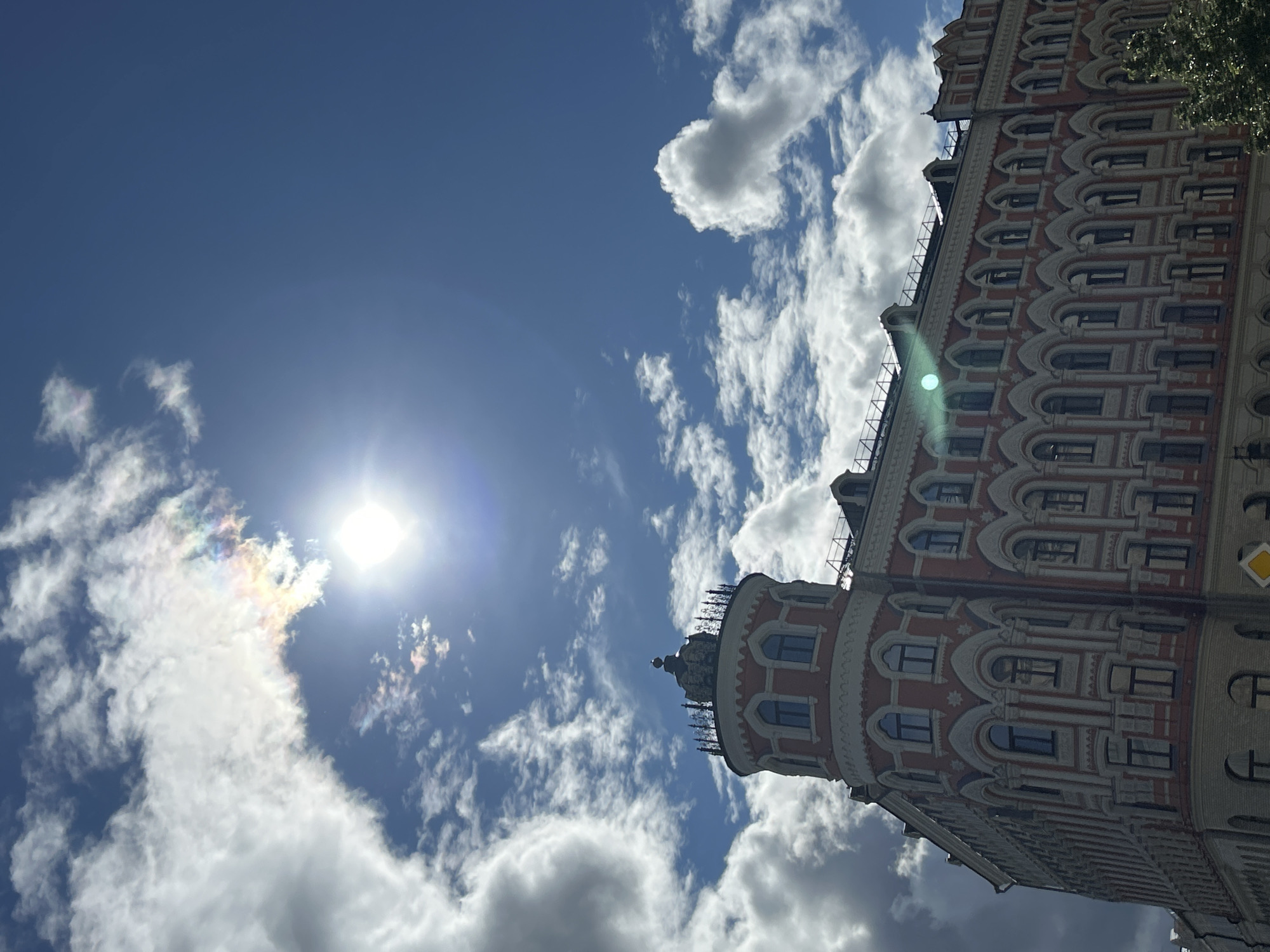 A low-angle shot depicts a building with ornate architectural details under a partly cloudy sky with the sun visible. The building is predominantly red with white trim, featuring arched windows and intricate patterns on its facade. A cylindrical tower-like structure with a darker roof and decorative elements is attached to the main building. The sky is bright blue with puffy white clouds, and the sun is positioned near the center of the frame, creating a bright glare.