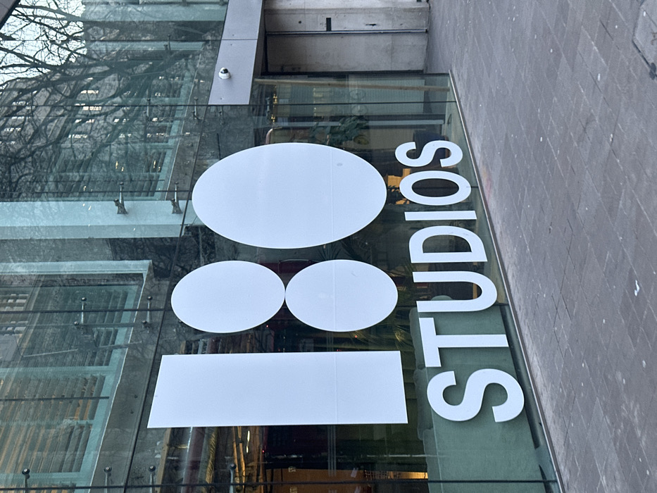 A close-up, angled shot shows the entrance to '18 Studios.' A large oval above two adjacent smaller ovals and a rectangle beneath them form the number 18. 'STUDIOS' is spelled out vertically in white letters on the right. The signage is on a glass facade reflecting a street scene. A security camera is visible above the entryway. The sidewalk is on the right side of the image.