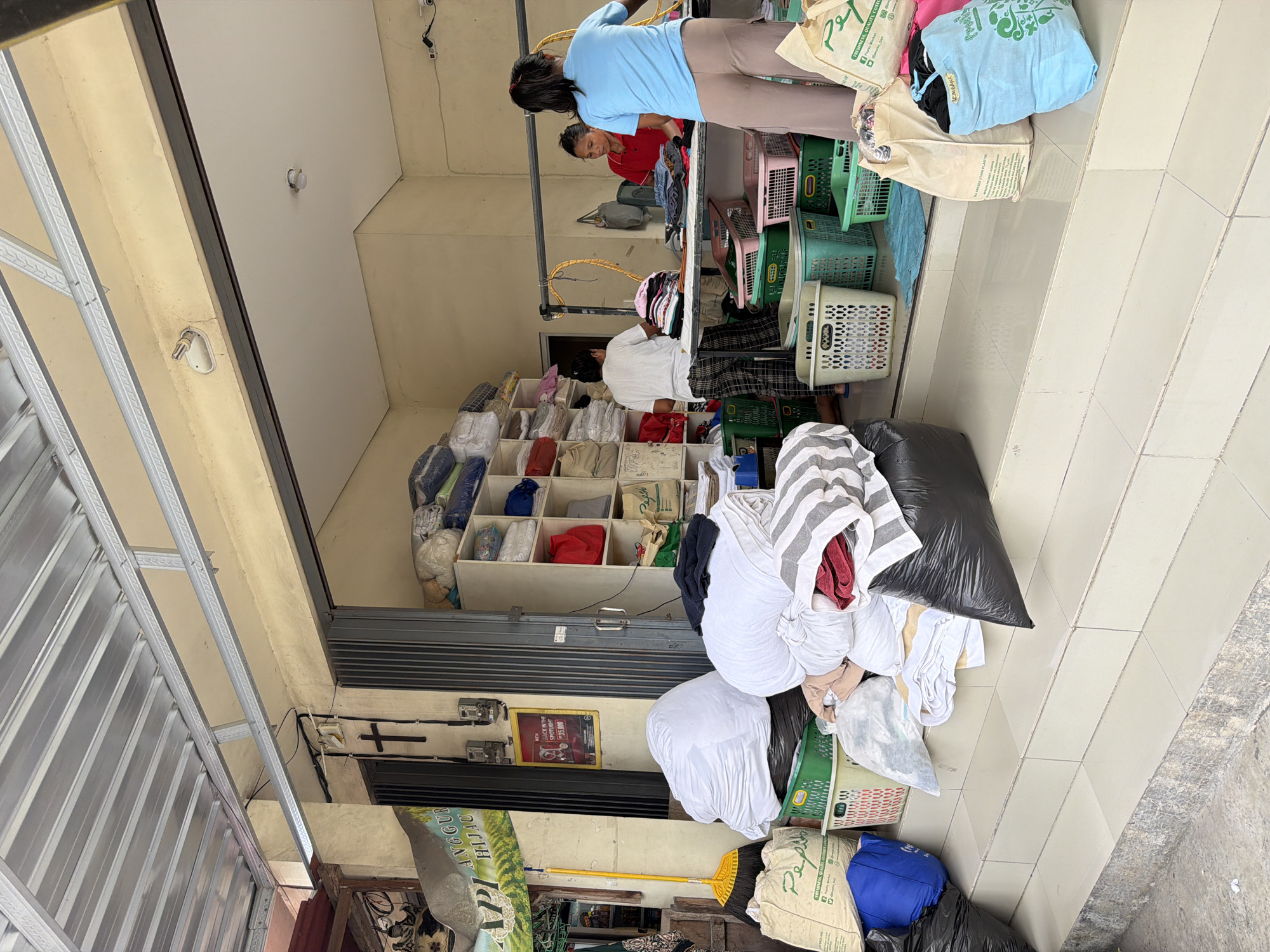 Here's a forensic description of the image:
**Scene:** A laundromat or dry cleaning business located in North Kuta, Indonesia.
**People:** Three individuals are visible. Two women are working near the front of the store, one wearing a blue shirt and one wearing a red shirt. Further back in the shop, another individual is present, dressed in white shirt and patterned pants.
**Objects:** Numerous items related to laundry services are present, including:
* Stacks of neatly folded clothes in shelving units.
* Large piles of unfolded laundry, some in black plastic bags, laundry baskets, and fabric bags.
* Fabric bags with green printing on them, including "Pegi."
* Clothes hanging on a rack.
**Environment:** The scene is inside a shop with a tiled floor. A sign containing the phrase "ANGGUR HIJAU" is visible.