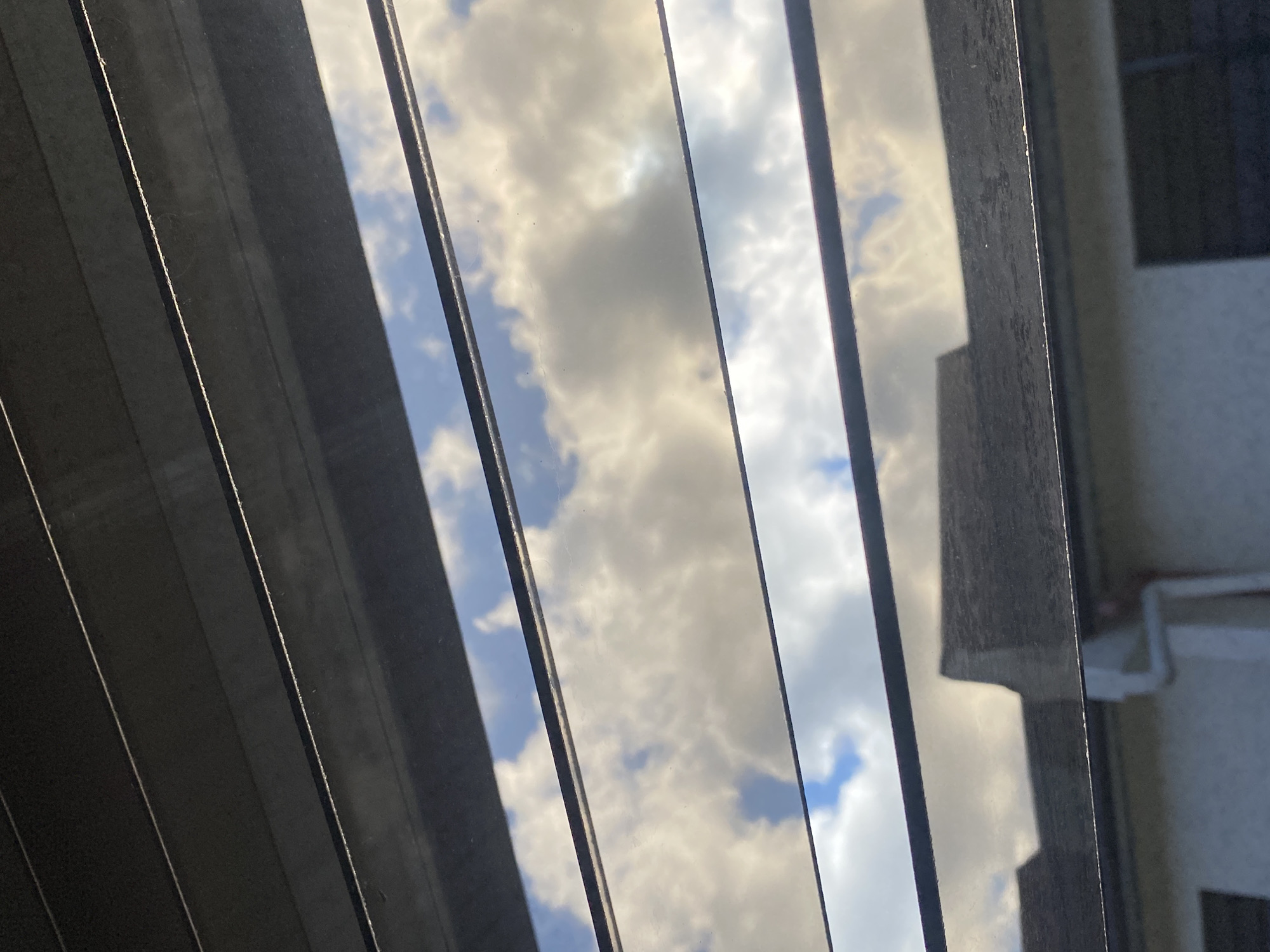 A view of the sky with white and grey clouds, partially obstructed by horizontal dark grey blinds. Below the blinds, a portion of a dark roof and a light-colored building is visible. There is a white gutter running down the side of the building. This image was taken in Davao City, Philippines.