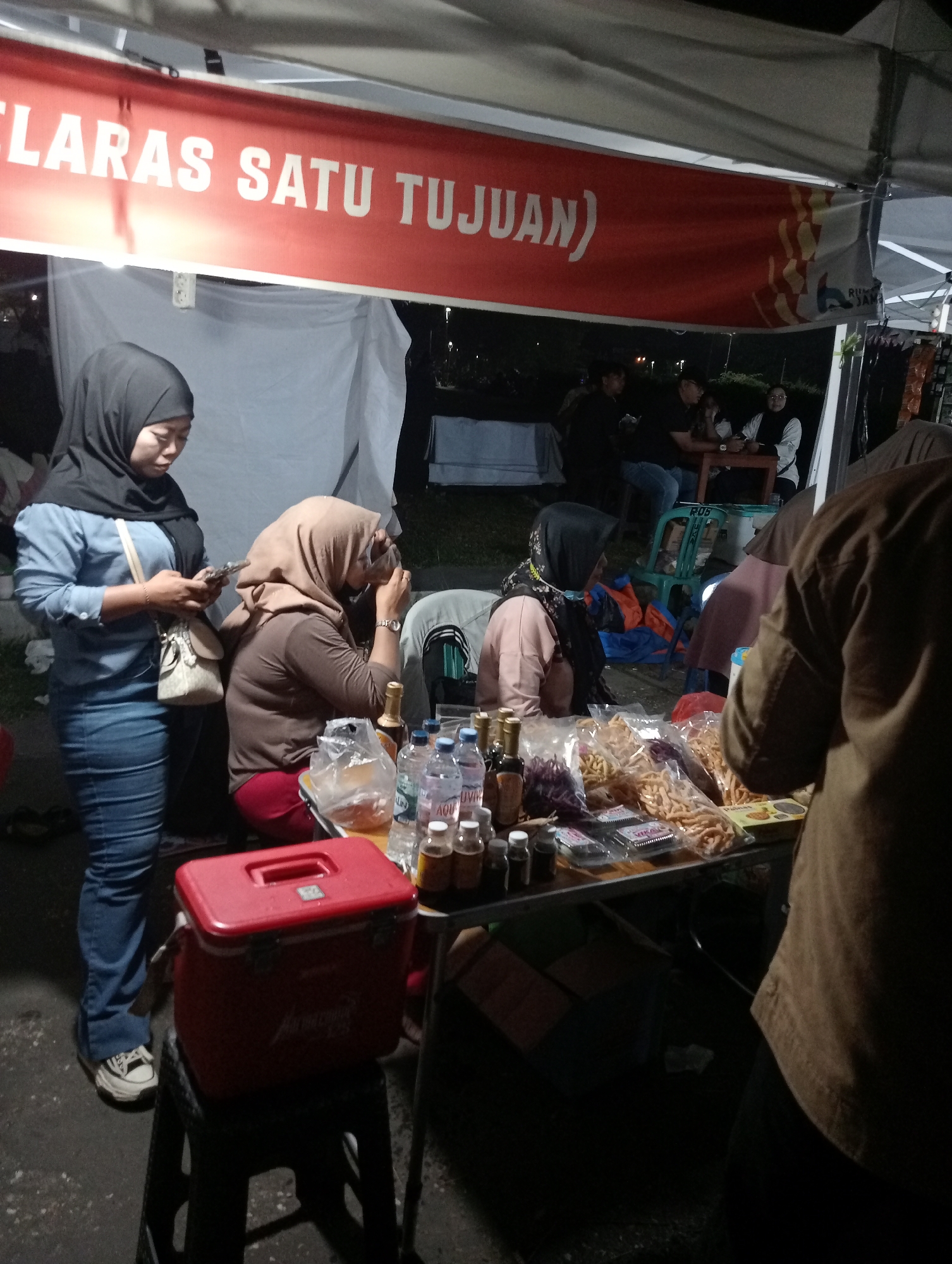 In Jambi, Indonesia, at night, a group of people are gathered around a food stall. A red banner with white text reads "KLARAS SATU TUJUAN". A woman in a blue shirt and jeans stands to the left, holding a phone and a bag. Another woman in a hijab and brown top is seated, looking down. In front of them, a table is laden with various snacks in plastic bags, bottles of water, and small bottles of what appear to be condiments. A red cooler box sits on a black stool. In the background, other people are seated at a table, and some are standing. The scene is illuminated by artificial lighting, creating a warm, ambient glow. The ground is dark and appears to be asphalt.