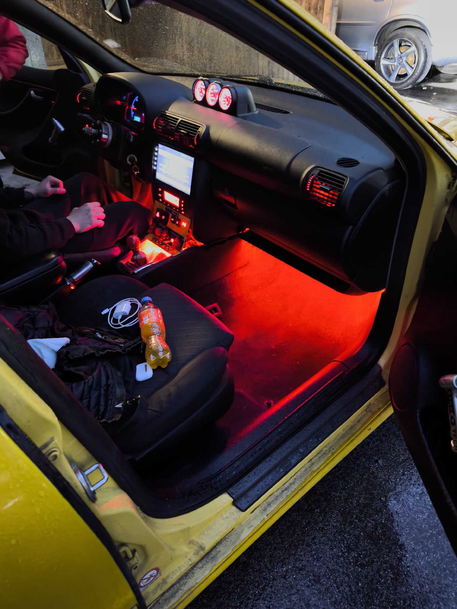 The image captures the interior of a yellow car, viewed from an open driver's side door, set in Burgas, Bulgaria. A person, likely male, is seated in the driver's seat, wearing dark clothing.
The car's interior is distinctly illuminated with a vibrant red ambient light, particularly noticeable in the footwells and emanating from the air vents. The main instrument cluster glows with blue light, while the aftermarket infotainment screen displays a lighter, likely white or blue, illumination. Three aftermarket gauges, also glowing red, are mounted on top of the dashboard.
On the passenger seat, an orange soda bottle, a white charging cable with a wall adapter, and white AirPods are visible, resting on a dark jacket or bag. The gear shifter area shows a customized setup, with a red element and a visible "Sparco" label on what appears to be a mat or trim piece. A silver car is partially visible outside in the background, past the open door.
The ground outside the car is wet, suggesting recent rain or washing. The time of day appears to be dusk or night, given the strong reliance on the car's interior lighting. The door jamb displays standard "no smoking" and airbag warning symbols. The overall scene depicts a customized car interior with a striking red lighting theme.